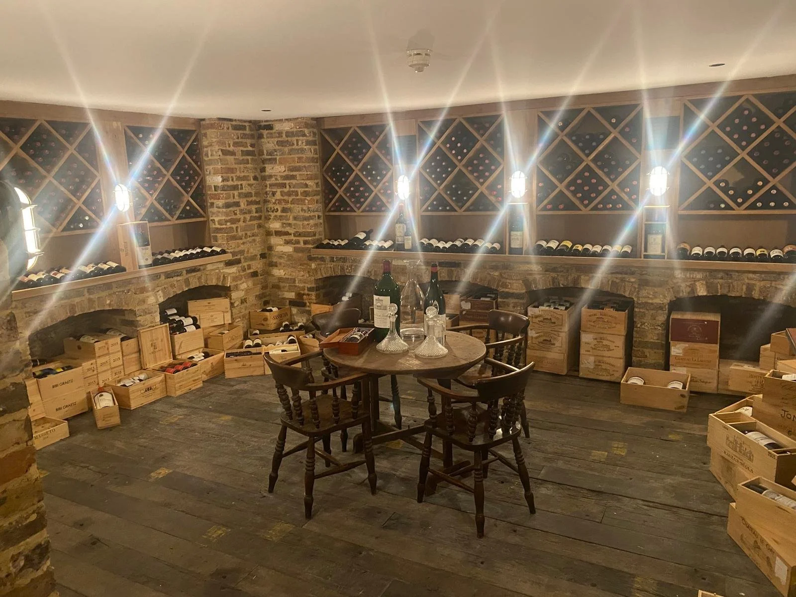 Wine cellar with wooden shelves filled with wine bottles, wine boxes on the floor, a round wooden table with four chairs, and a concrete and brick wall background.