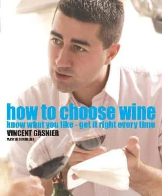 A man holding a glass of red wine, with a wine book titled 'How to Choose Wine' in the background.