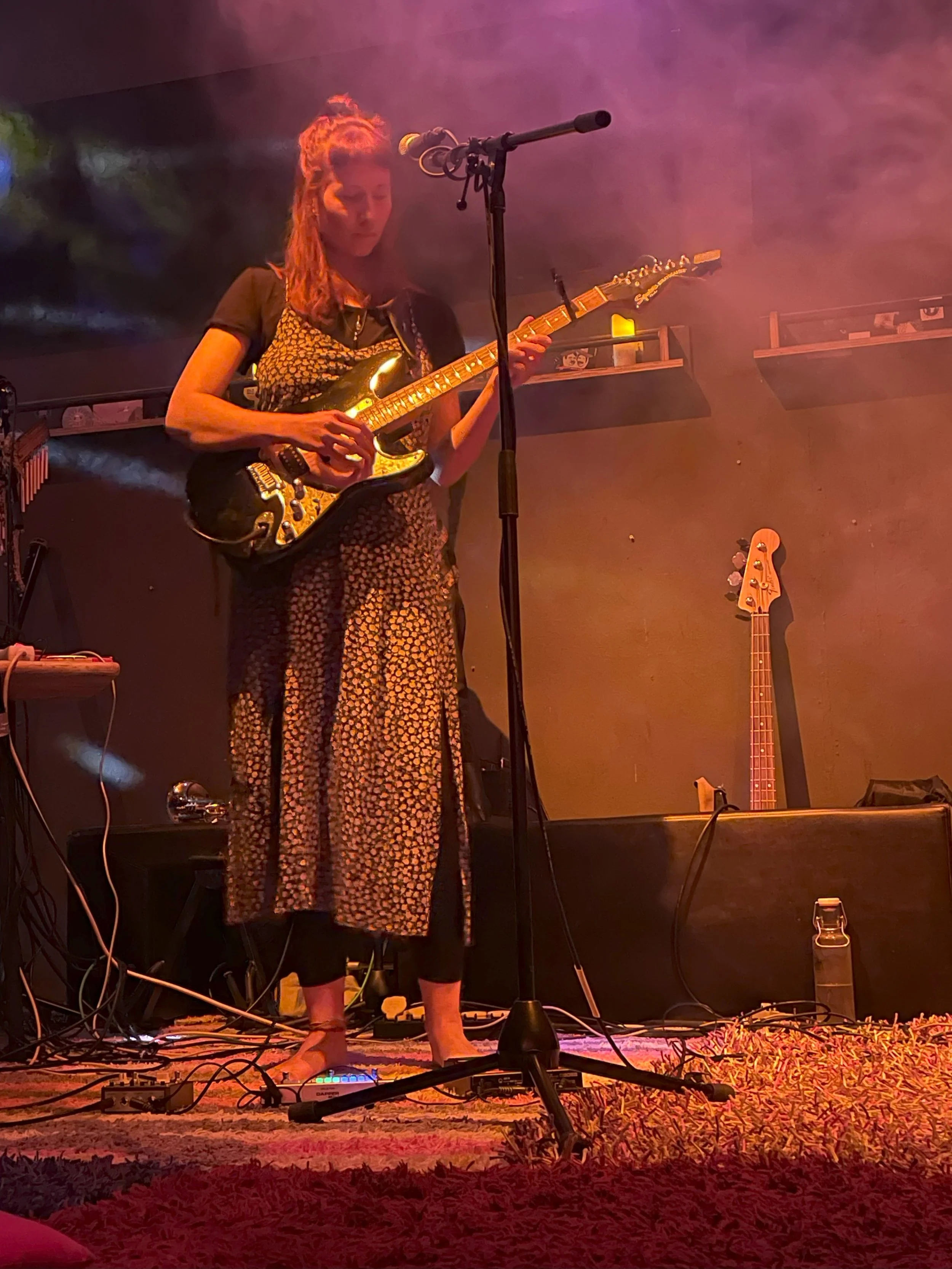 Eine Frau spielt E-Gitarre auf einer Bühne mit Mikrofon und Effekten, umgeben von buntem Licht und Nebel.