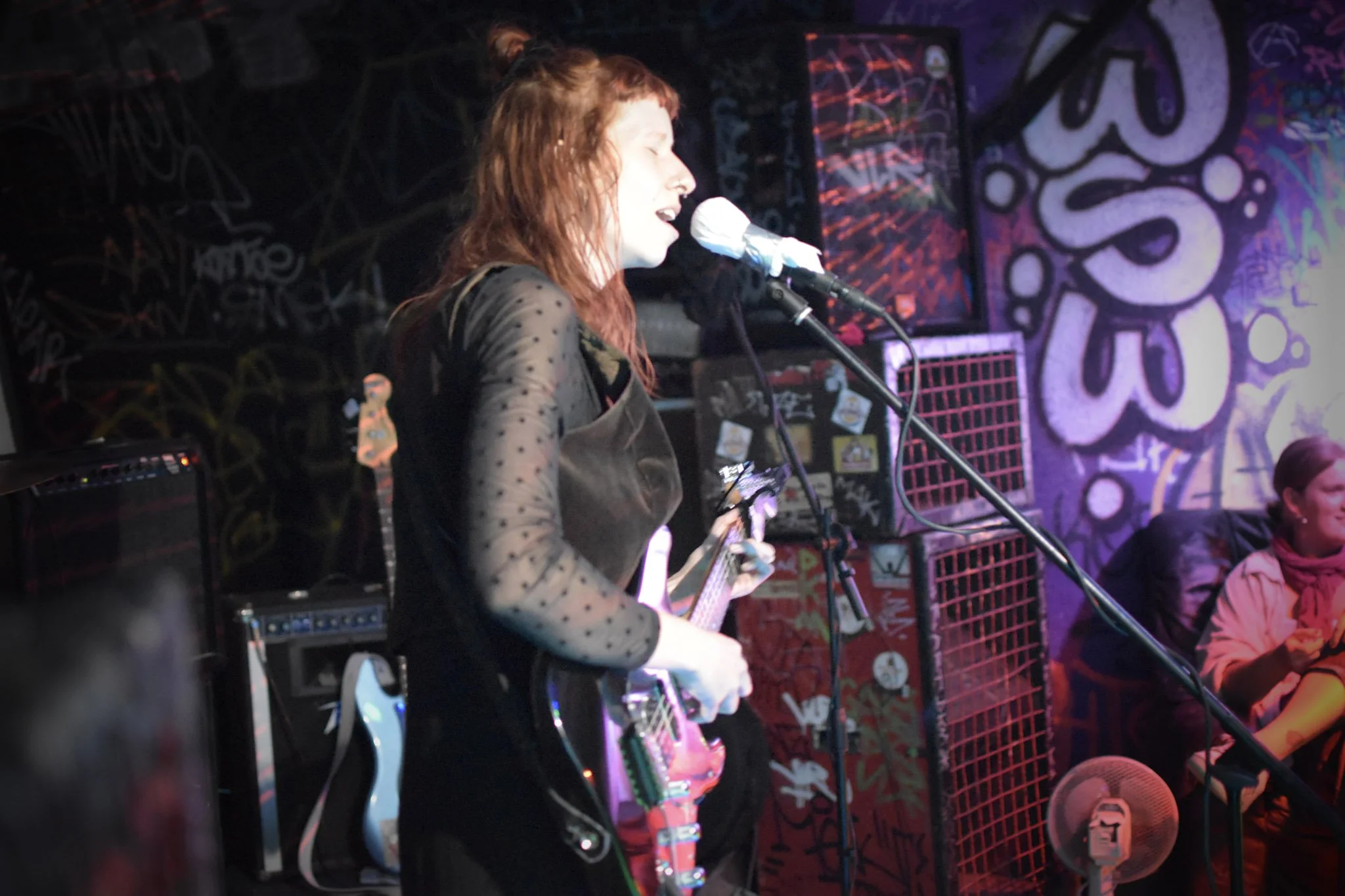 Frau spielt E-Gitarre und singt vor Mikrofon in einem Club, Wände mit Graffiti verziert, weitere Person im Hintergrund sichtbar.