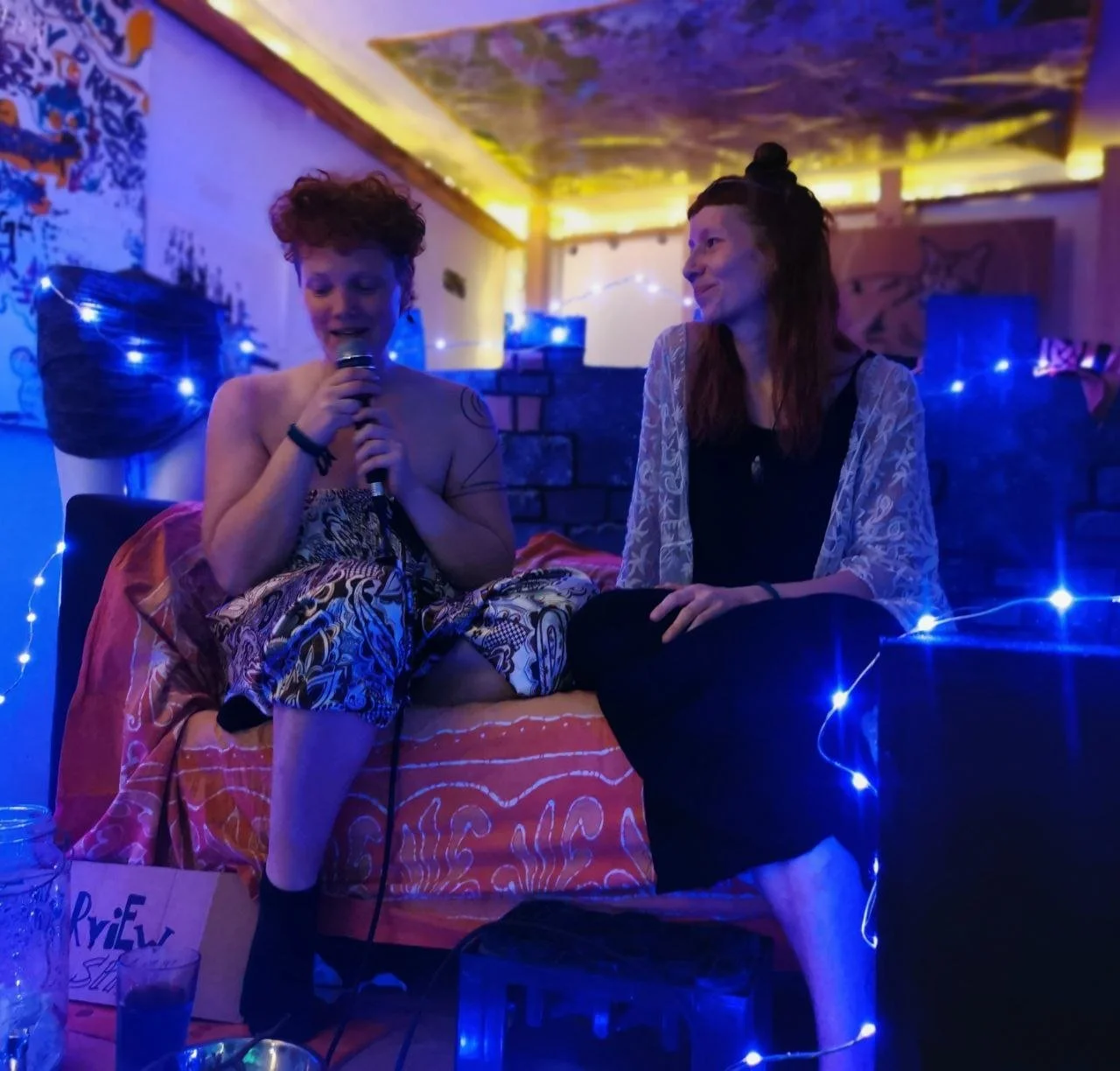 Zwei Personen sitzen auf einem Sofa, umgeben von blauer Beleuchtung und dekorativen Lichtern. Die Person links hält ein Mikrofon, während die Person rechts lächelt. Im Hintergrund sind bunte Wanddekorationen sichtbar.