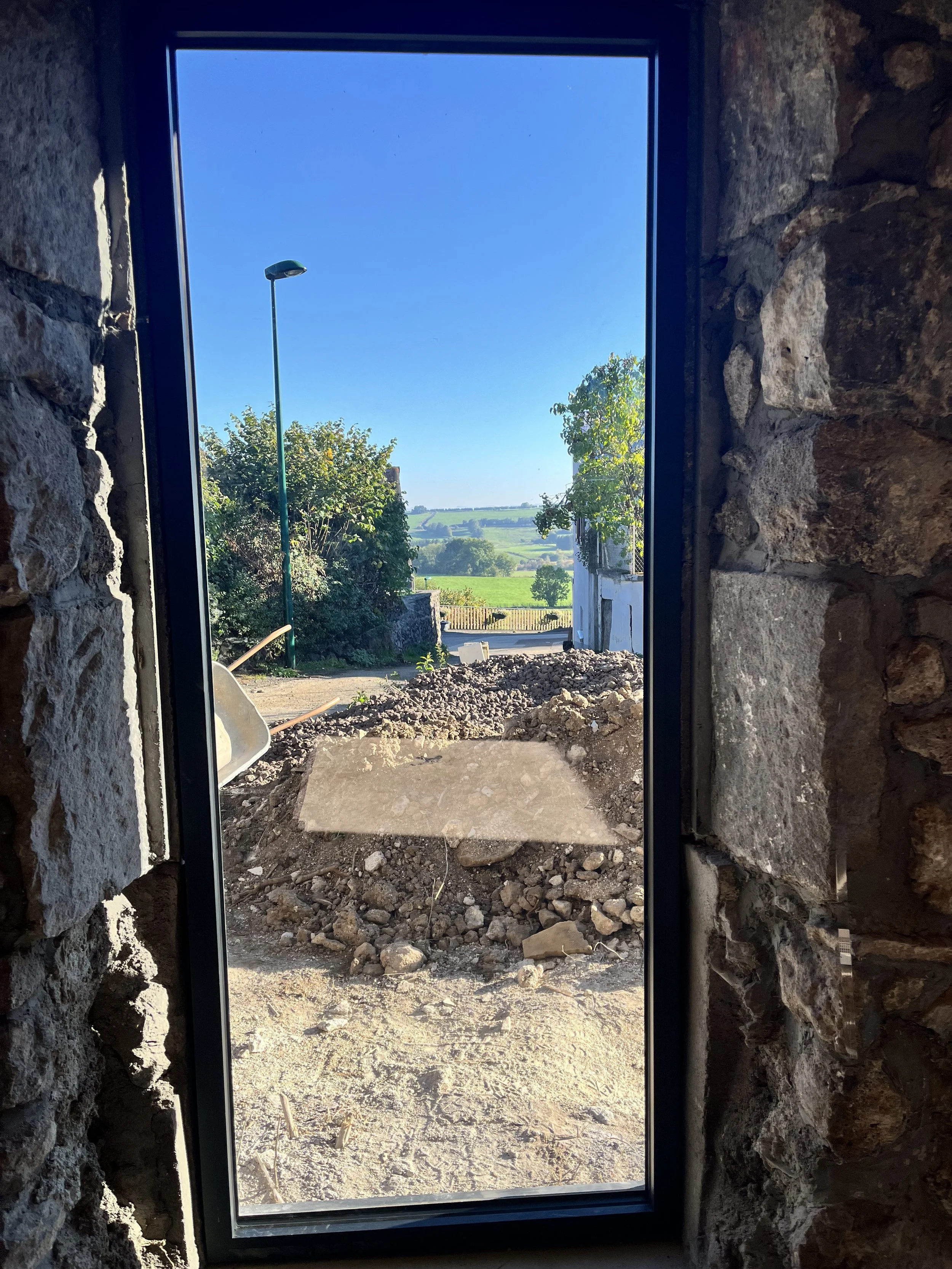 Vue extérieure à travers une fenêtre en verre montrant un chemin en brique avec des piles de terre et un lampadaire, avec des arbres, des champs verts et un ciel bleu clair.