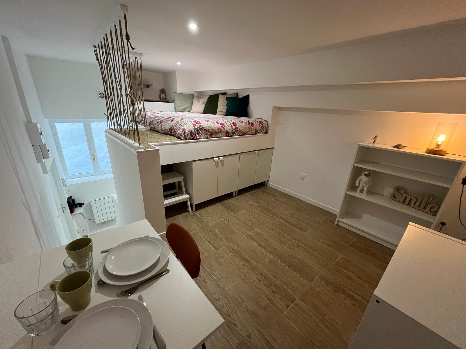 Intérieur d'un appartement avec un lit mezzanine, une table à manger avec des assiettes et des verres, une étagère décorative, et des lumières douces.