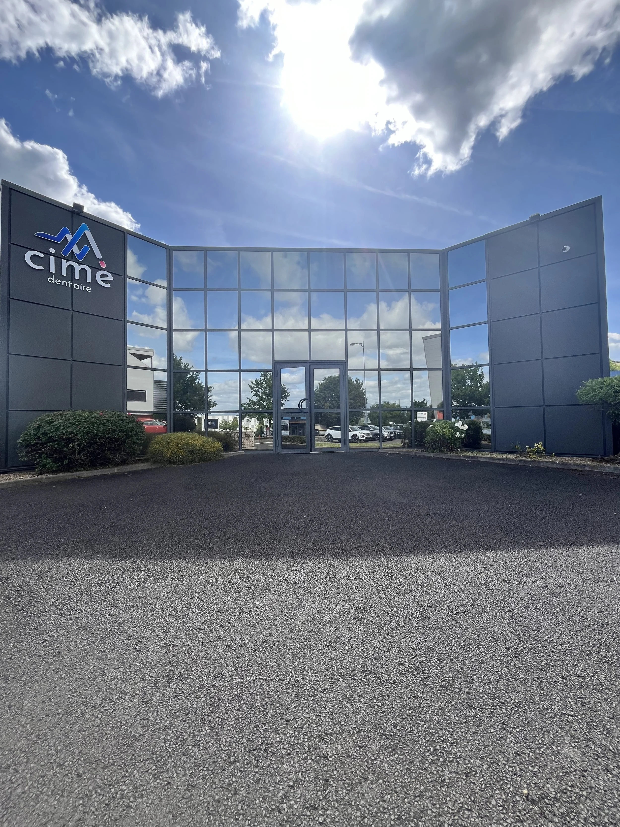 Bâtiment moderne avec une façade en verre et un logo 'cime dentaire' sur le côté gauche, sous un ciel en partie nuageux avec le soleil brillant.