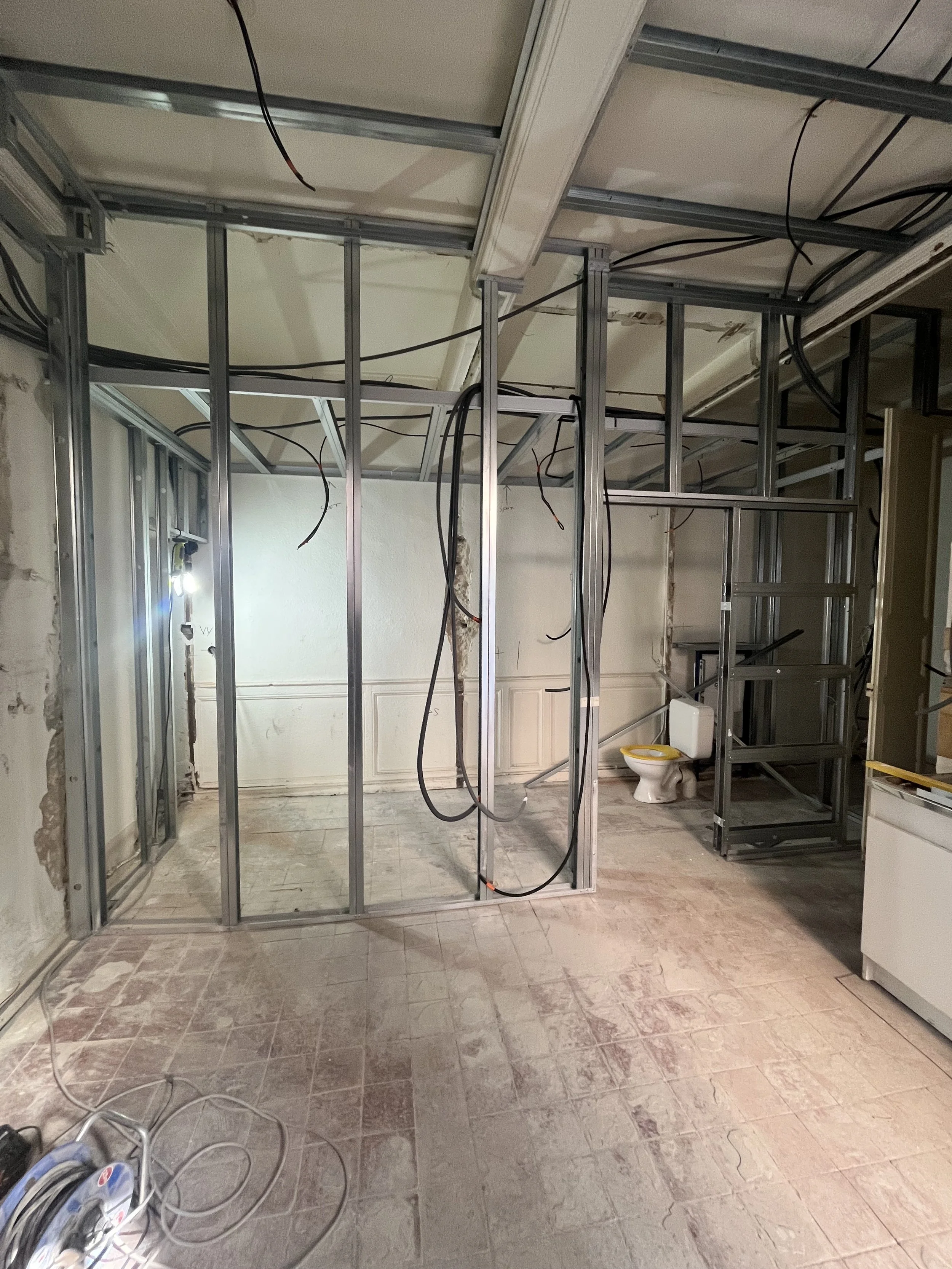 Une pièce en rénovation avec une structure métallique pour un mur ou un plafond, des câbles électriques suspendus, un lavabo de toilette, et un sol en carreaux en cours de nettoyage ou de pose.