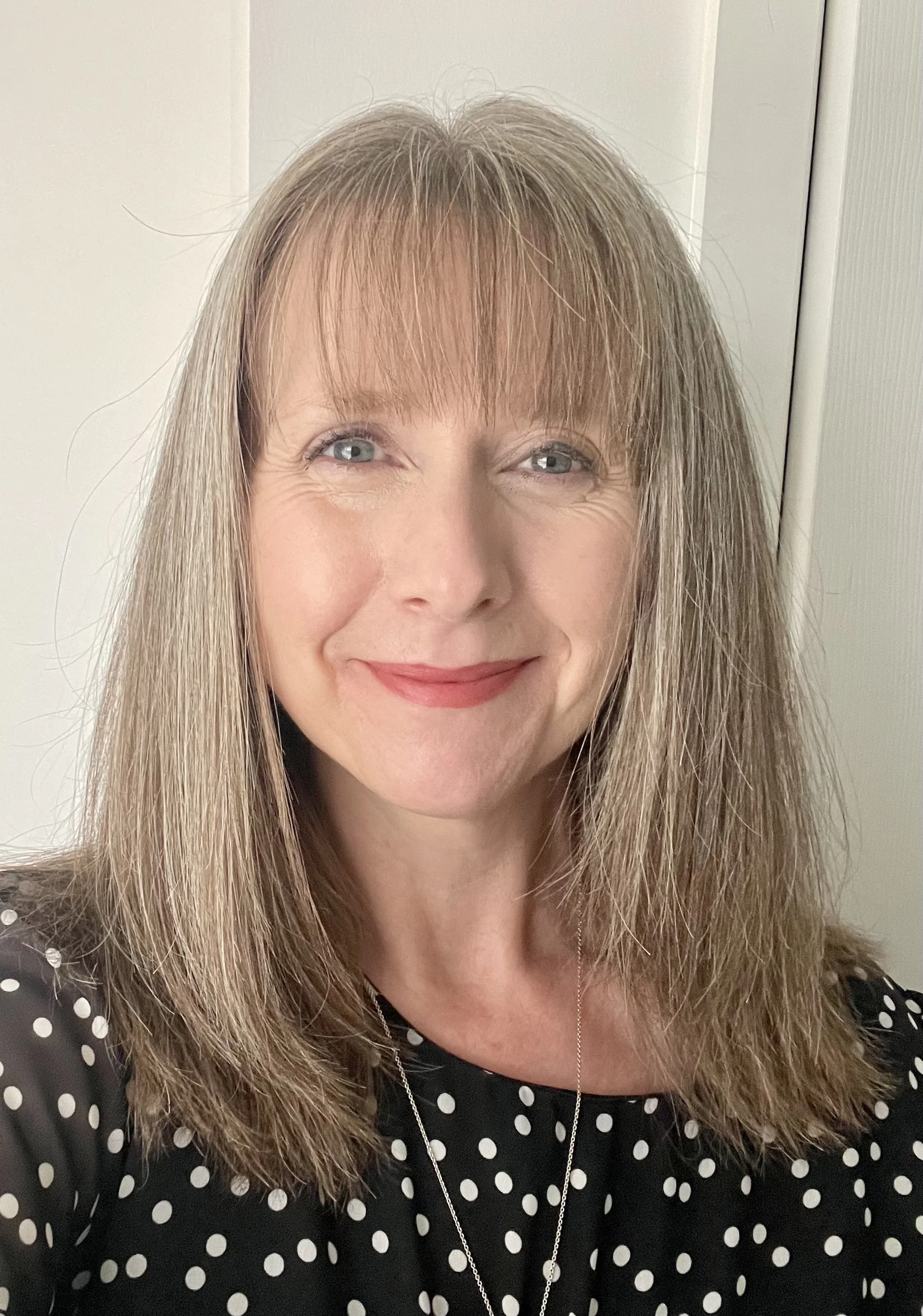 A middle aged woman with medium length brown and grey hair and blue eyes who is smiling and wearing a black and white spotty top.