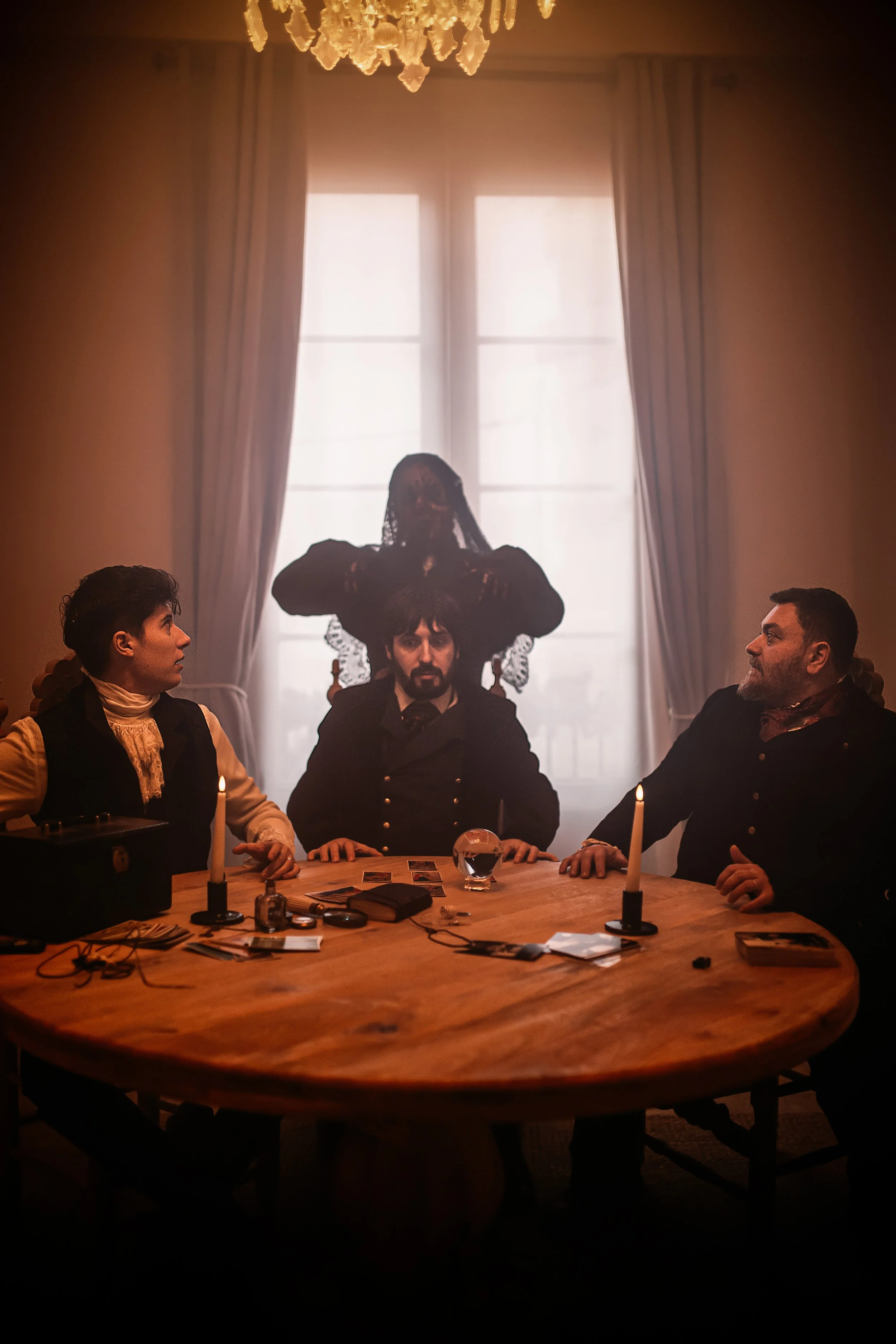 Tres personas sentadas en una mesa redonda de madera, con velas encendidas, y una mujer fantasmal con velo | ReVolution | Rol en Vivo España