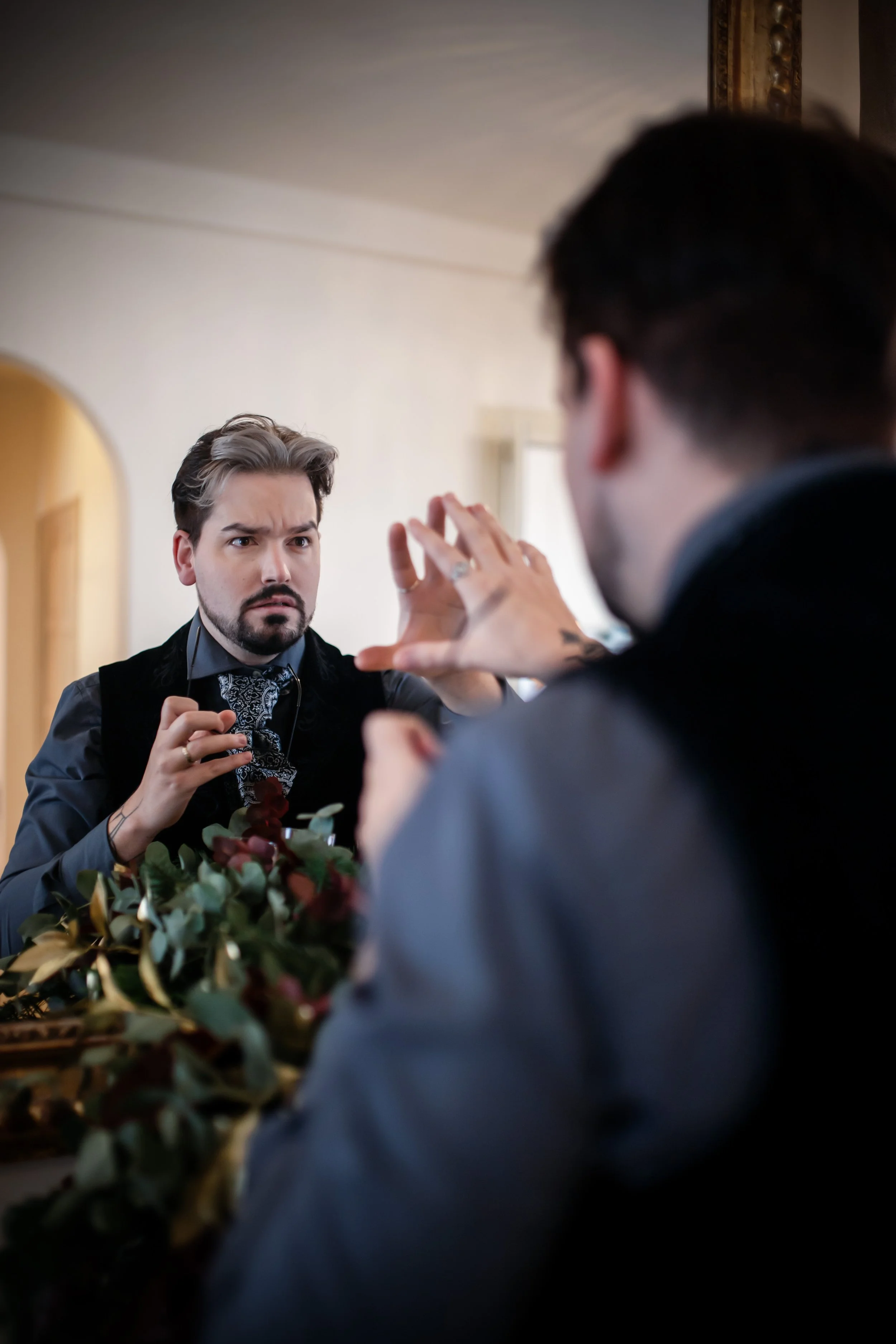 Hombre con barba, bigote, cabello peinado, vestido con chaqueta oscura, mirando con expresión de sorpresa o preocupación en un espejo | ReVolution | Rol en Vivo España