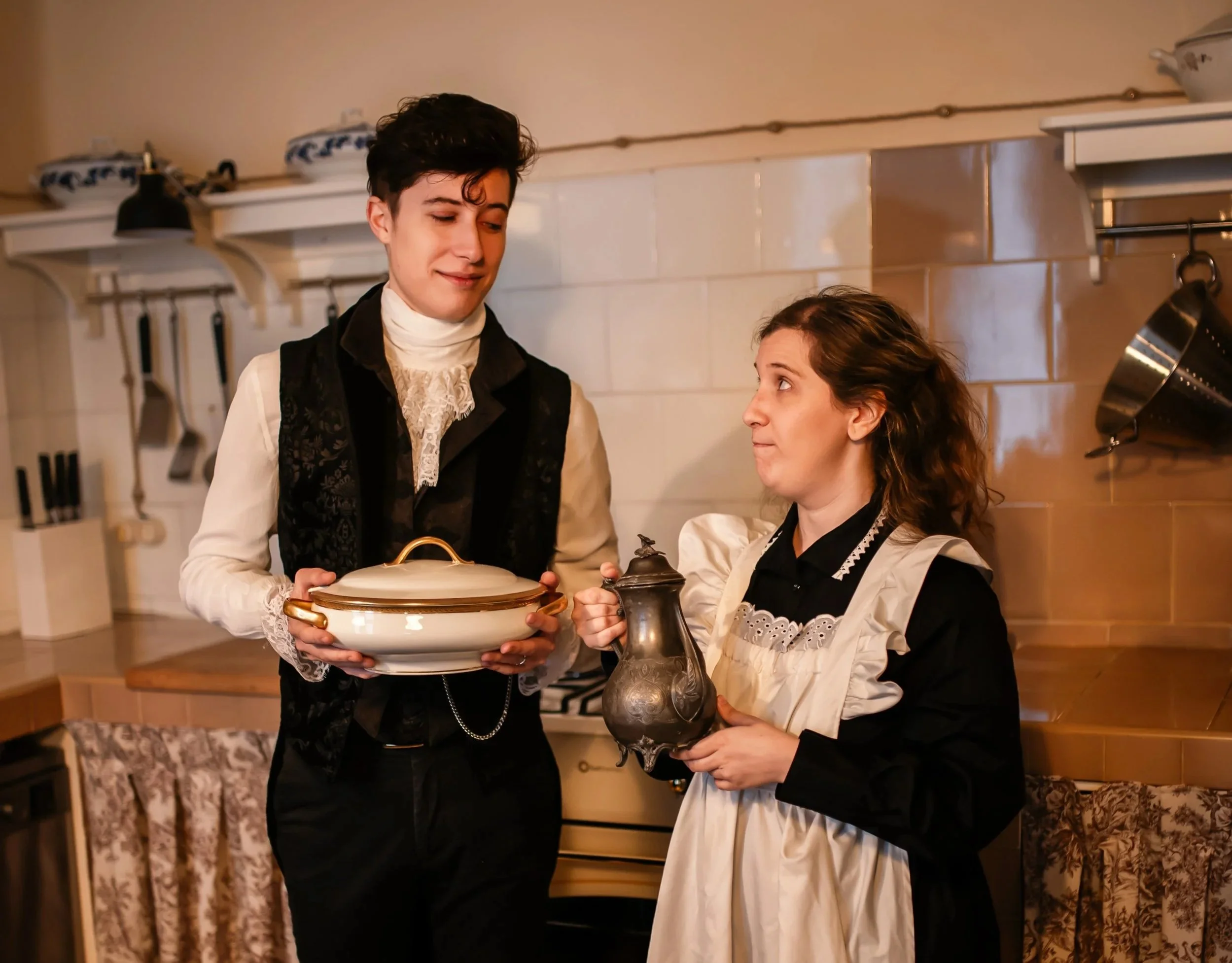 Una escena de época en una cocina, un joven con vestimenta antigua y una joven con delantal están intercambiando una vasija y una jarra de metal, respectivamente.