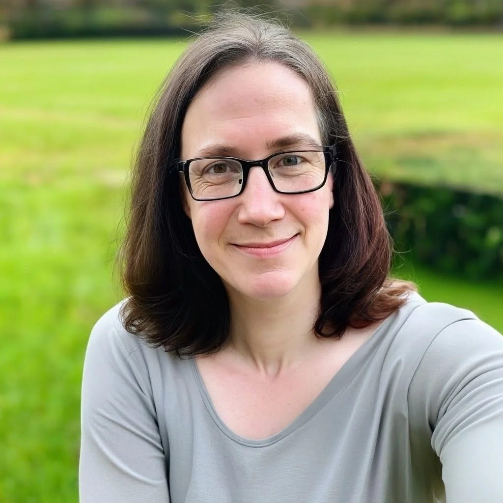 A woman with glasses and shoulder-length dark hair smiling outdoors, with a grassy field and trees in the background.