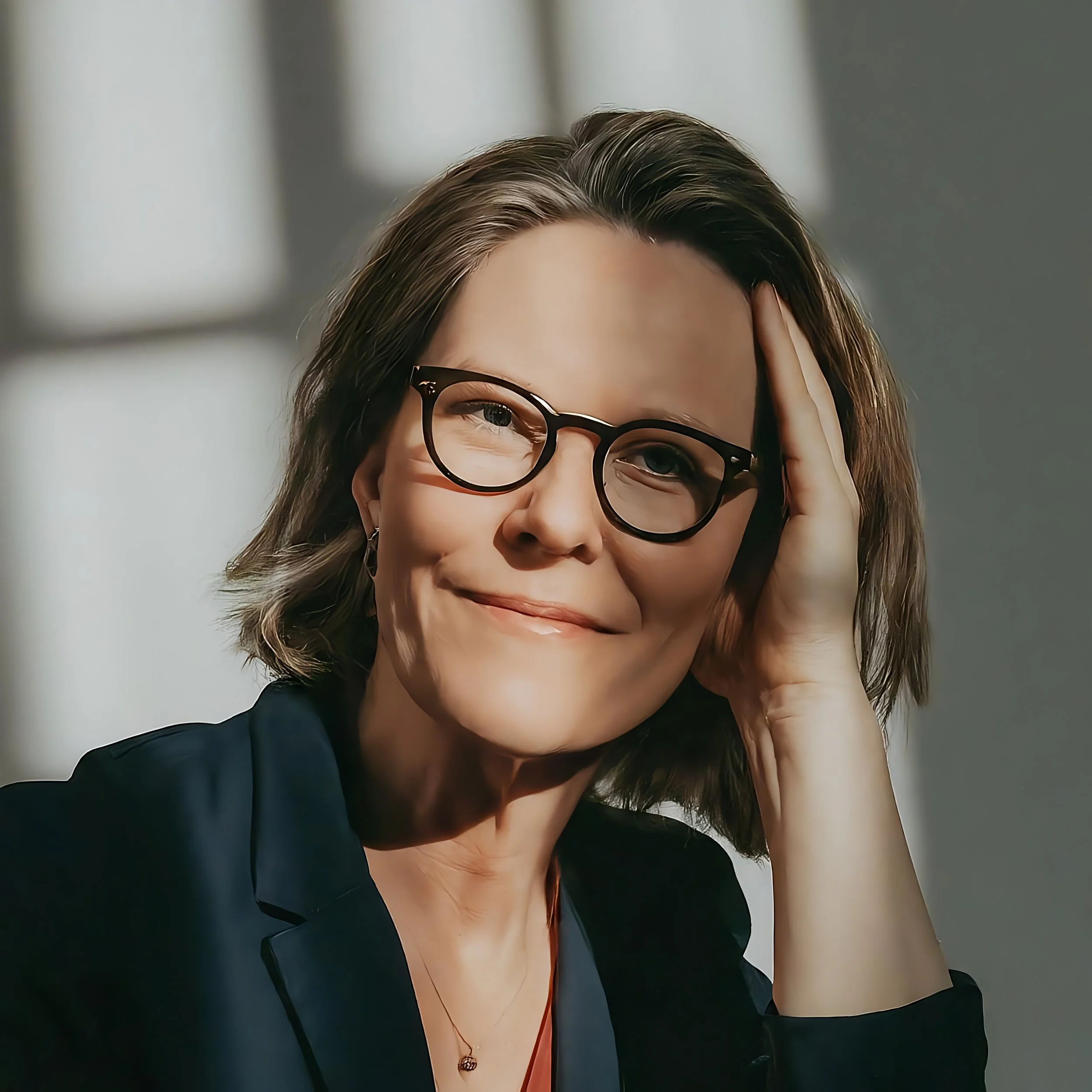 A woman with short brown hair wearing glasses, a black blazer, and a subtle smile, resting her head on her hand.