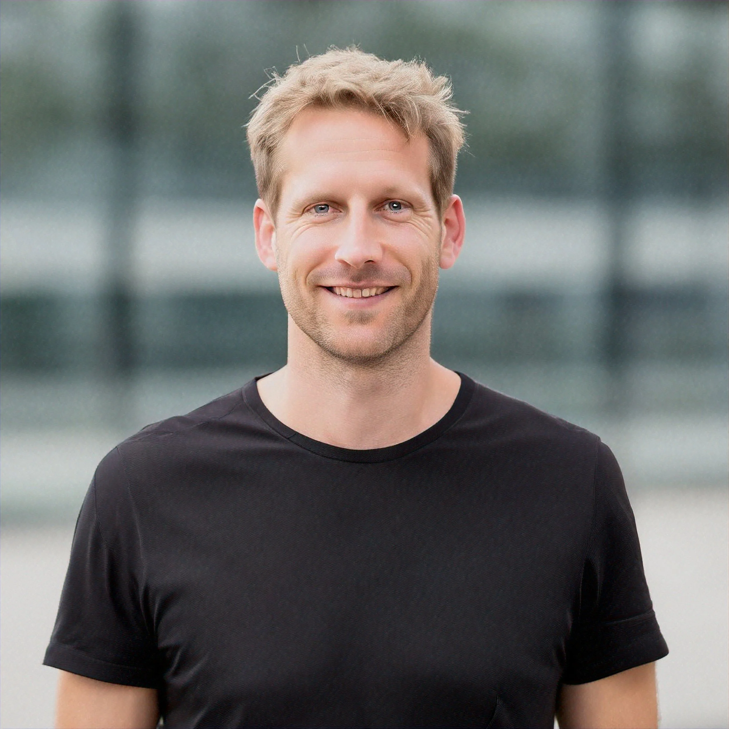 A smiling man with short blonde hair wearing a black t-shirt stands outdoors with a blurred background.