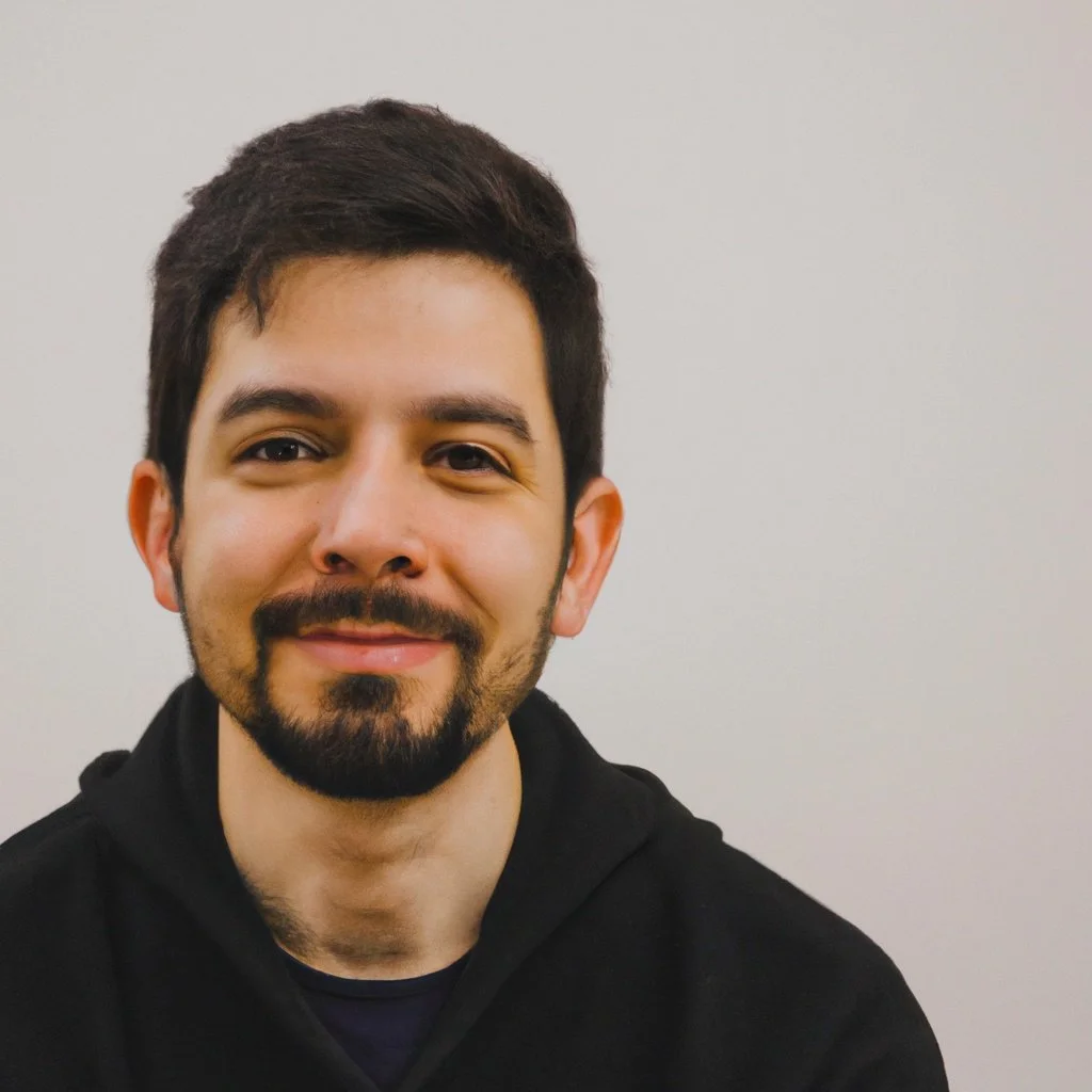 A man with dark hair and a beard, smiling, wearing a black hoodie, against a plain light-colored wall.