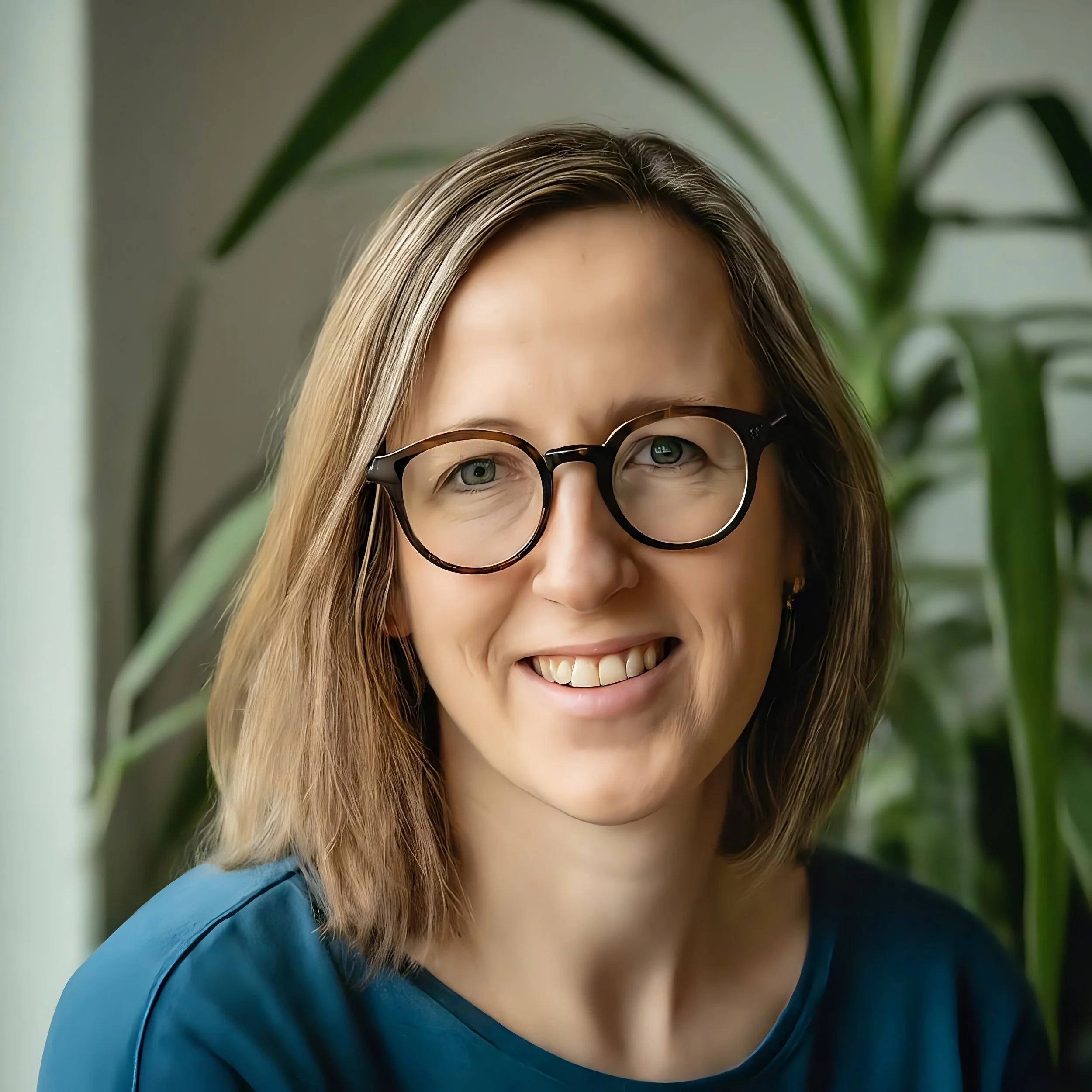 Close-up photo of a smiling woman with shoulder-length light brown hair, wearing black-rimmed glasses and a dark blue top, with large green plants in the background.