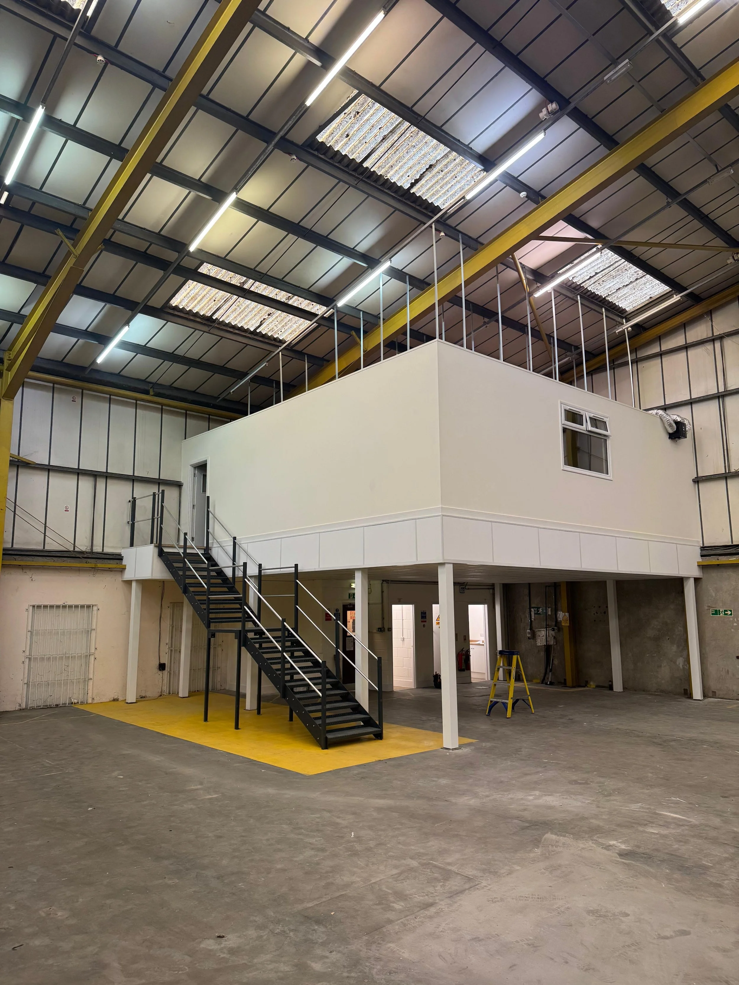 Interior of a large warehouse with a mezzanine office structure, black staircase, concrete floor, and high ceiling with skylights.