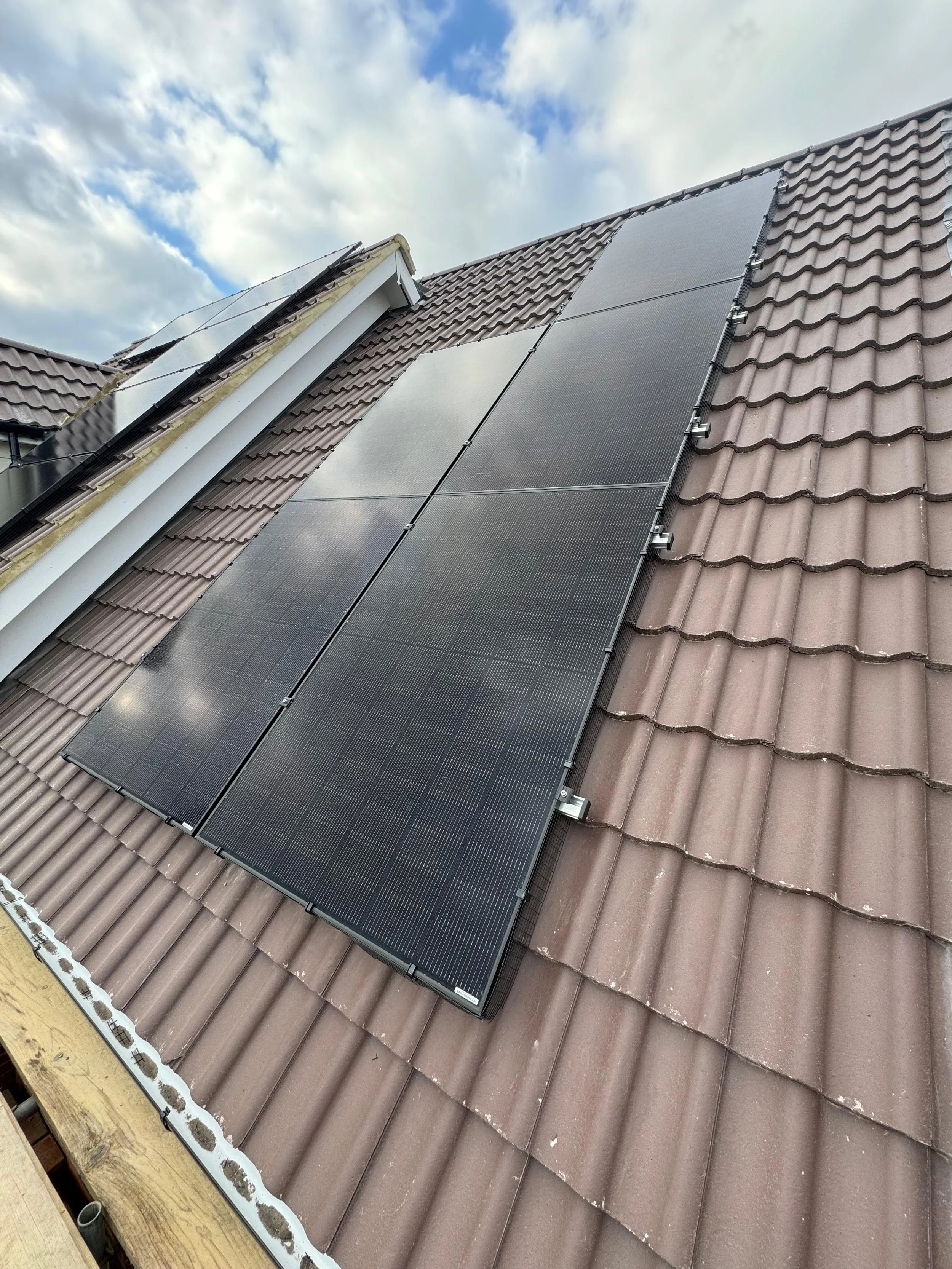 Black solar panels installed on a brown tiled roof under a partly cloudy sky.