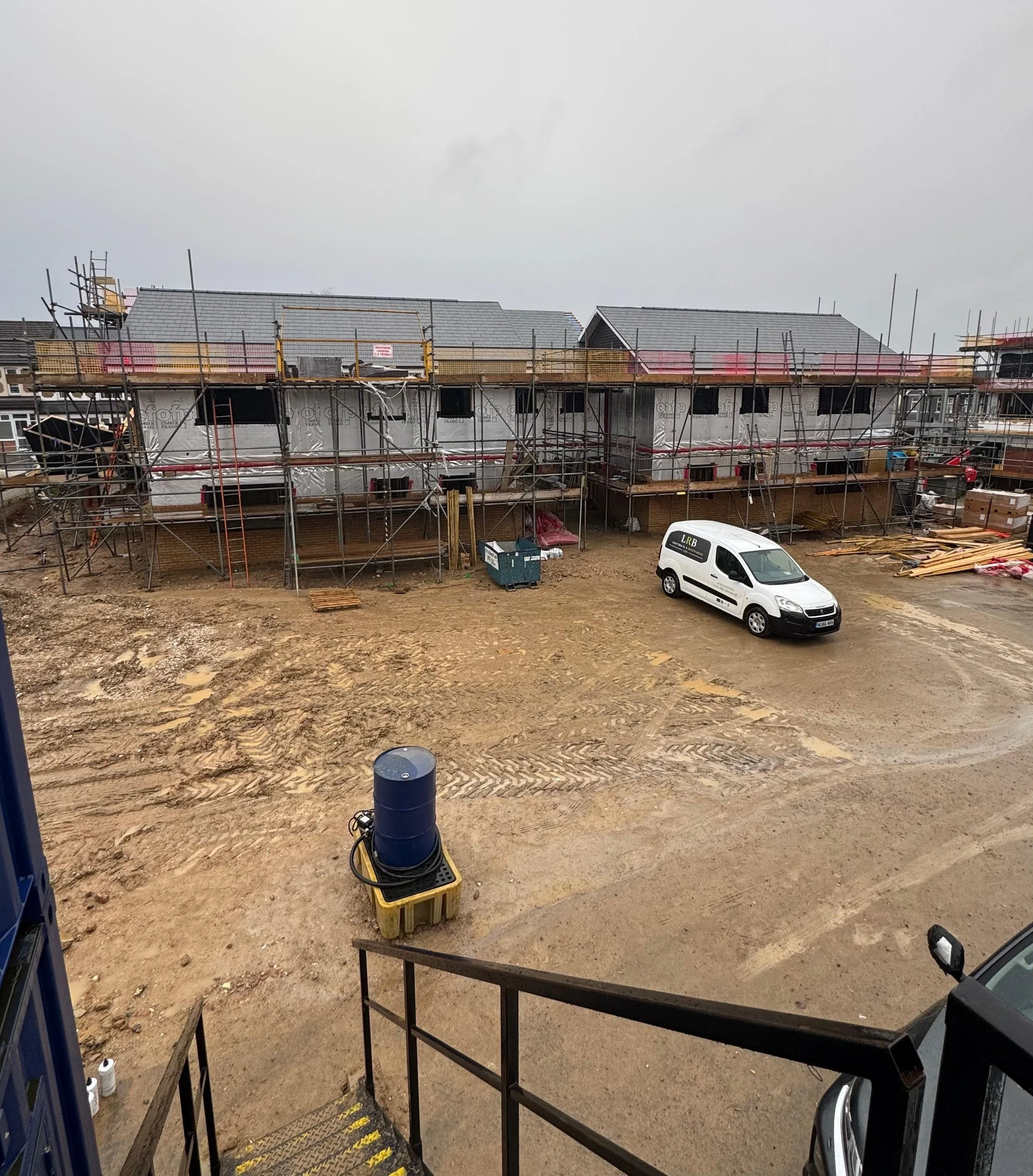 Construction site with scaffolding around new houses, parked white van, muddy ground, and overcast sky.