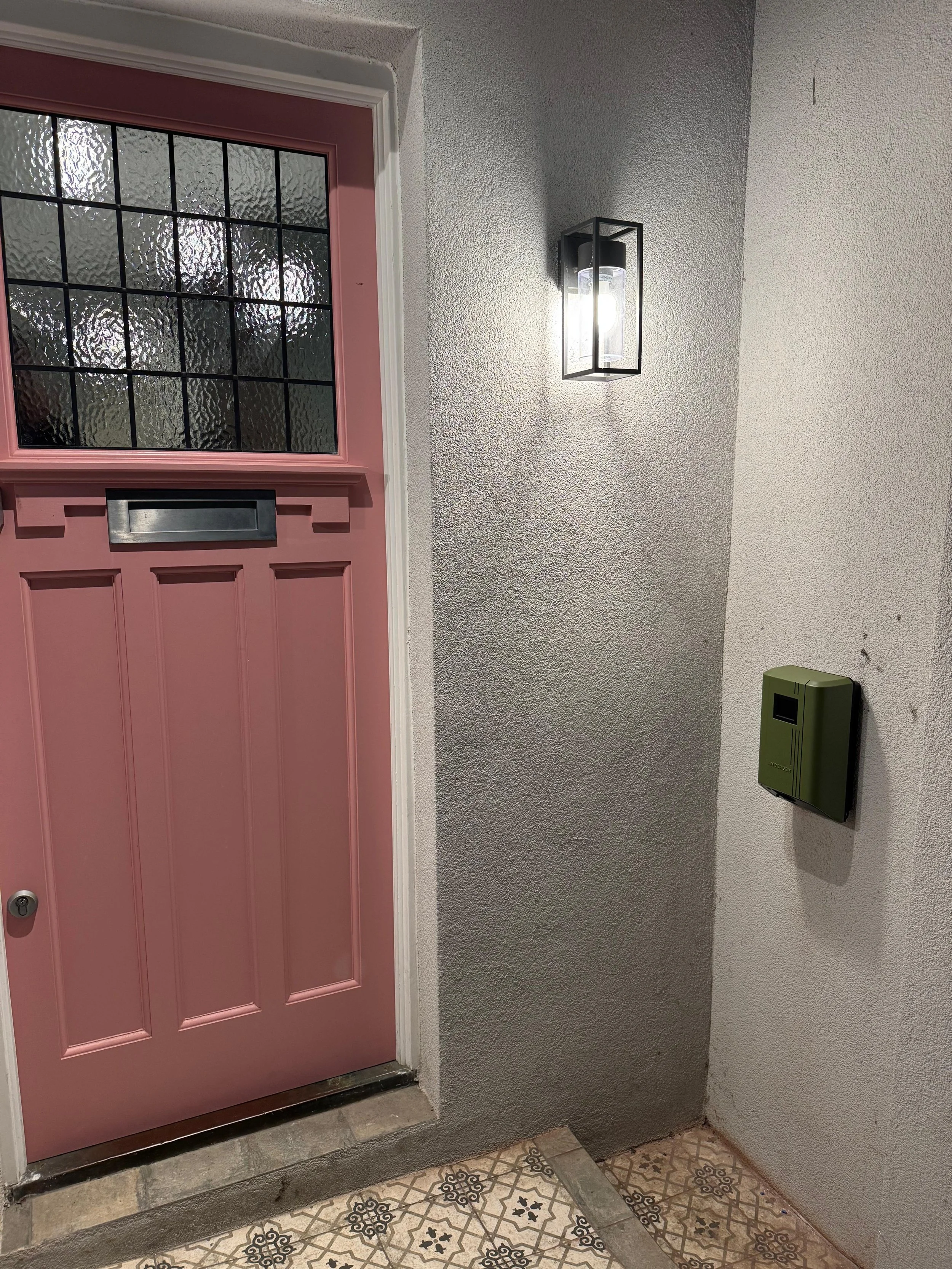 A pink front door with windowpanes on top, a mailbox slot, and a peephole. Next to the door, there is a wall-mounted green security panel. A modern wall sconce with a glass cover and black frame lights the area. The wall is textured gray, and patterned tiles cover the floor.