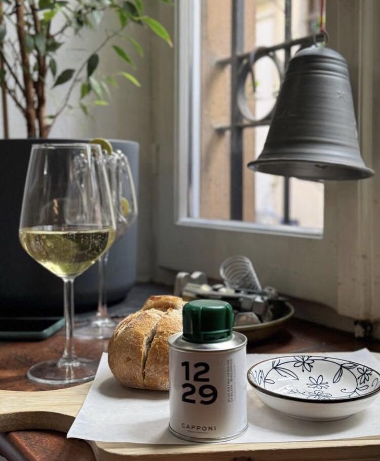 Scena su un tavolo con un bicchiere di vino bianco, pane, una lattina di olio d'oliva etichettata "1229 Capponi" e un piattino decorato. Sullo sfondo, un campanello appeso e una pianta vicino a una finestra.