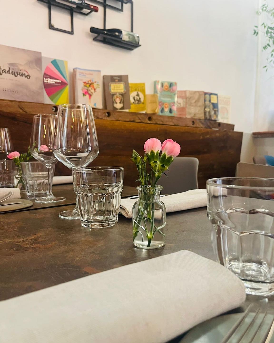 Tavolo apparecchiato con bicchieri e fiori rosa, mensola con libri sullo sfondo.