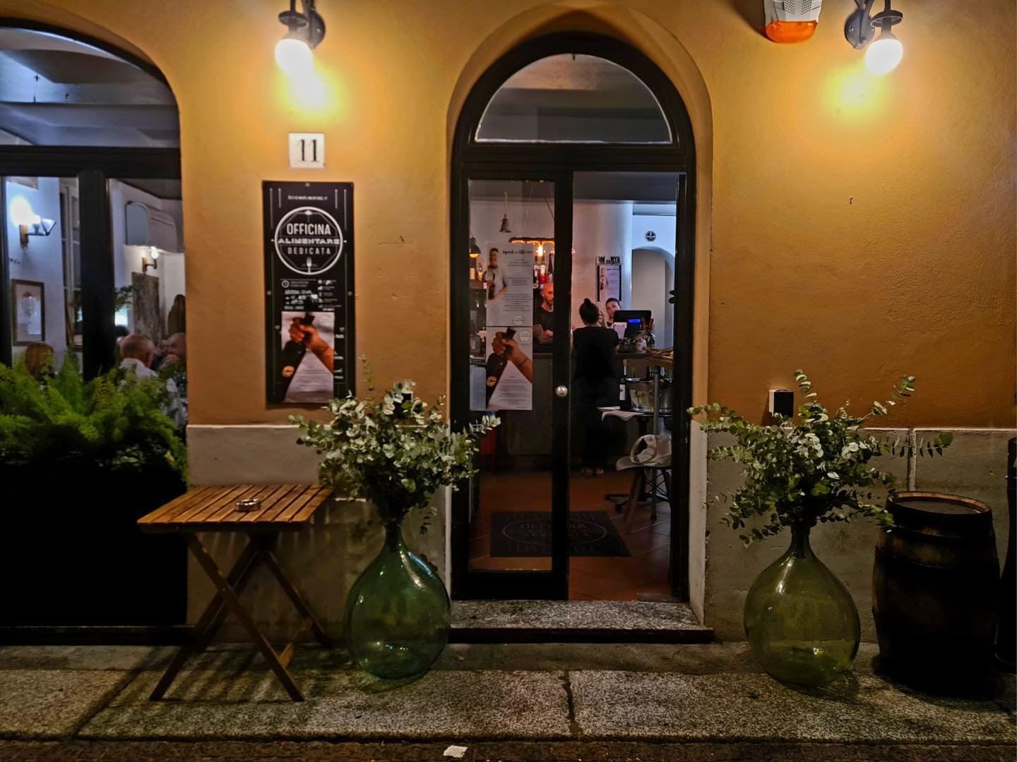 Ingresso di un ristorante con decorazioni e luci serali, tavolo in legno, vaso con piante e bidone, persone all'interno.