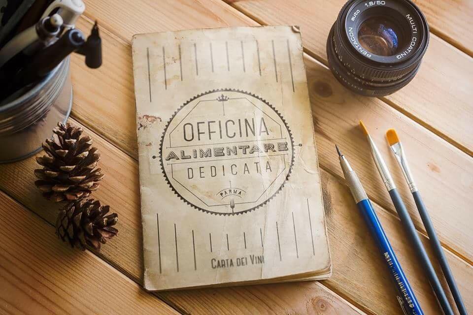 Tavola di legno con un libro intitolato "Officina Alimentare Dedicata", corredato di un obiettivo fotografico, un contenitore con penne e pennelli, e due pigne decorative.