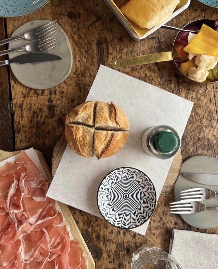 Tavolo apparecchiato con prosciutto crudo, pane rotondo, posate, piattino decorato e sott'aceti. Immagine di cibo italiano.