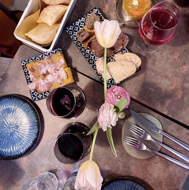 Tavolo apparecchiato con piatti, calici di vino rosso, petali di fiori, pane e salumi su piatti decorati.