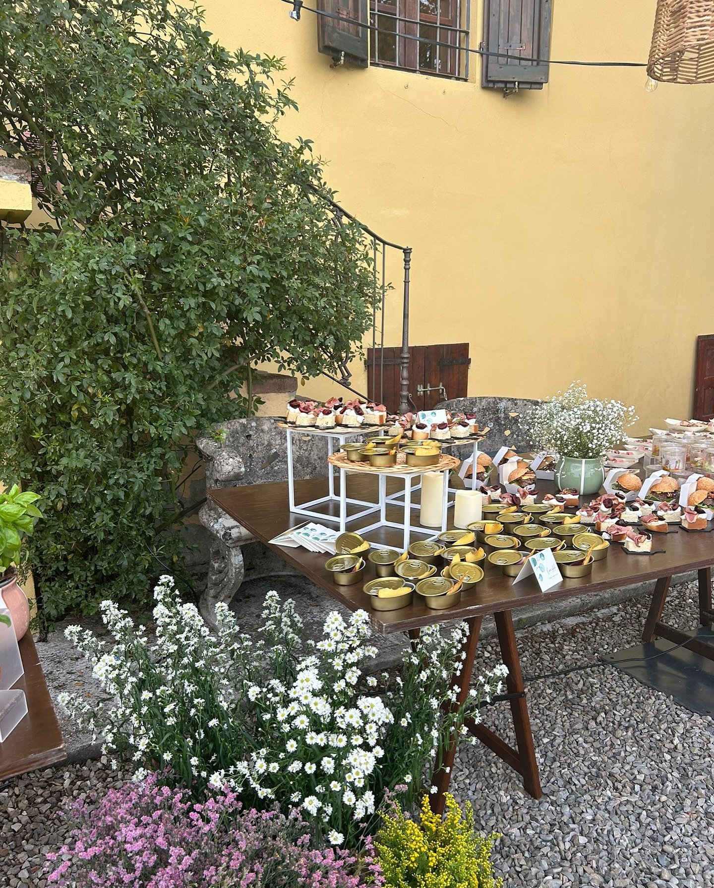 Buffet dolci all'aperto con tavolo marrone, vasi di fiori, e cespuglio verde vicino a una casa gialla.