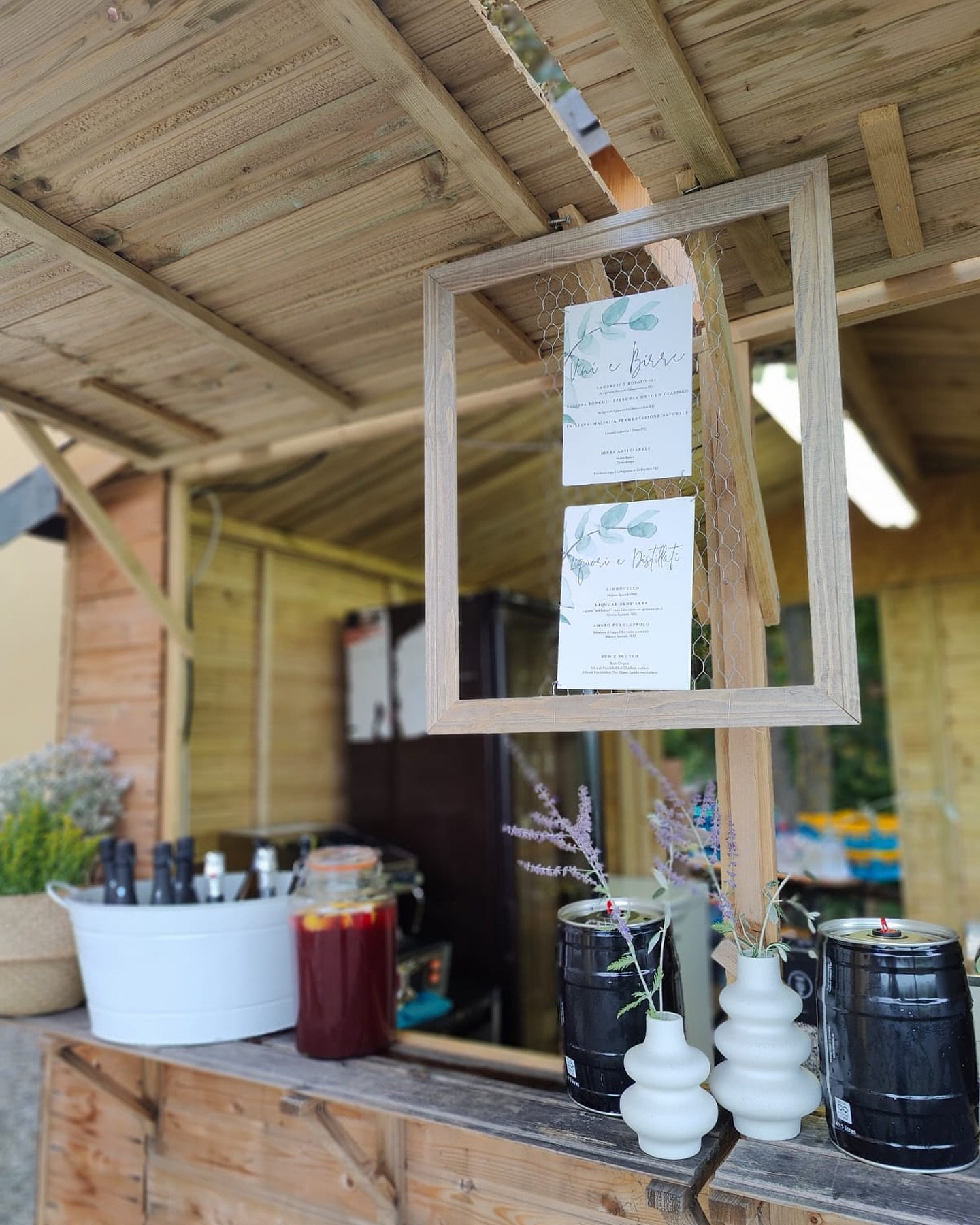 Un chiosco in legno con un menu appeso in una cornice di rete. Sul banco ci sono contenitori di bevande, vasi bianchi con fiori viola e un barile nero. Sul lato si vede un grosso contenitore con bottiglie.