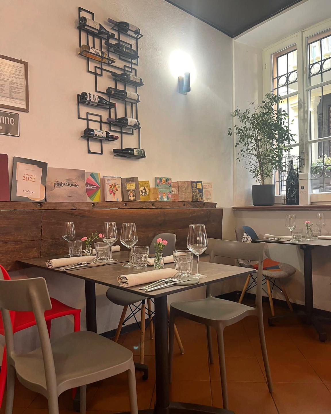 Interno di un ristorante elegante con tavoli apparecchiati, sedie colorate, una mensola con vini e libri, e una pianta vicino alla finestra.