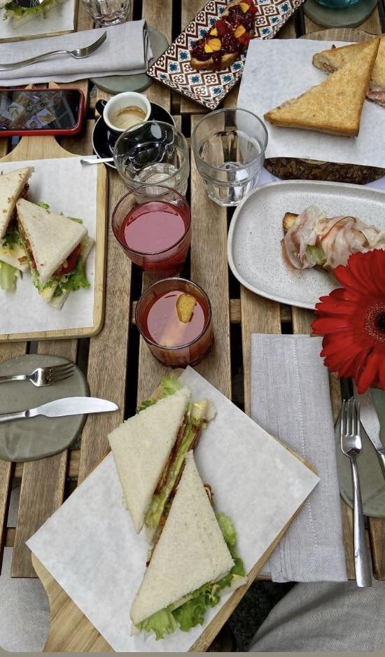 Colazione su un tavolo di legno con sandwich triangolari, affettati su un piatto, bicchieri di succo d'arancia, un caffè, una forchetta e un coltello su tovaglioli, e un telefono rosso.