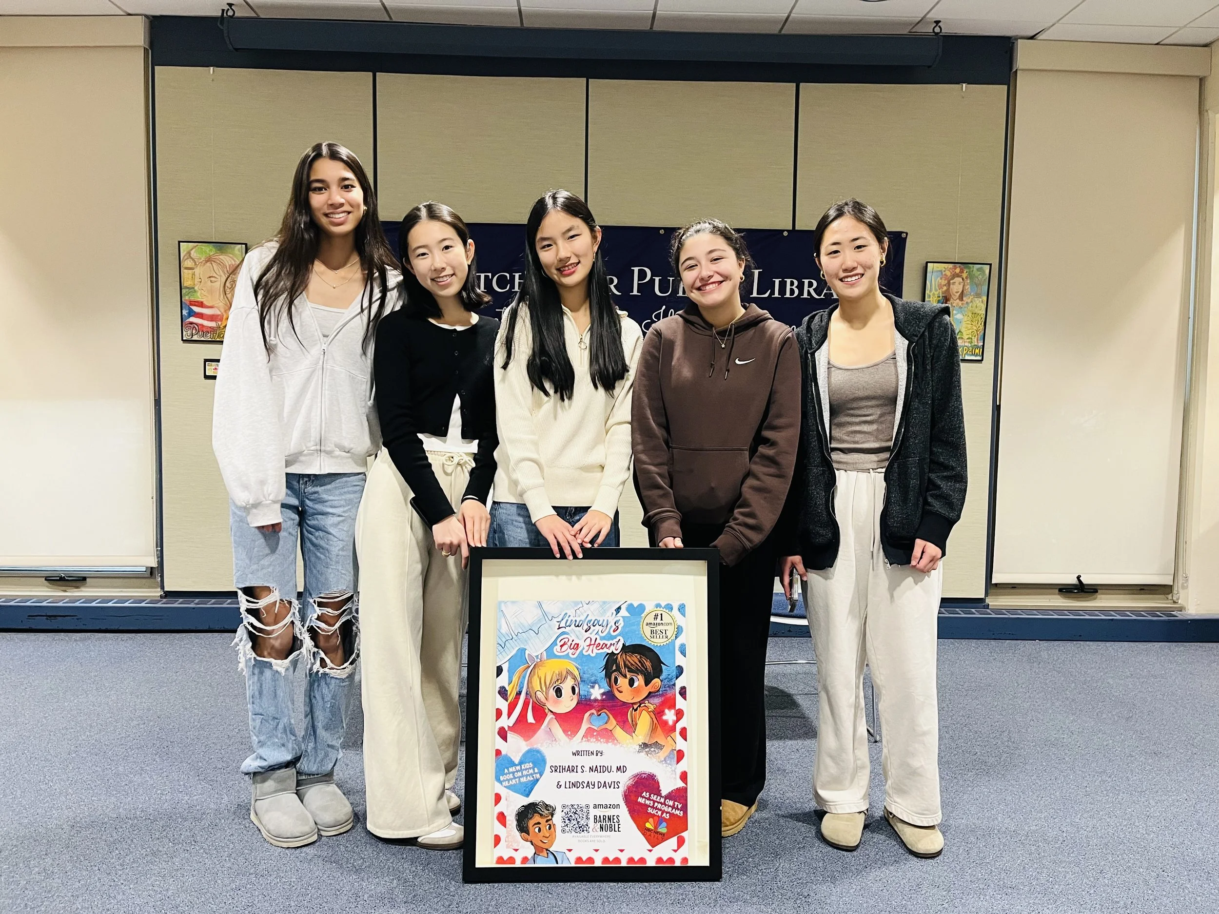 Six young women standing together in a library, smiling for a group photo. In front of them is a framed book cover titled 'Lindsay's Big Heart' written by Srihari S. Naidu, MD and Lindsay Davis, featuring colorful cartoon illustrations of a girl and a boy with hearts and stars in the background.