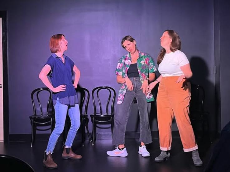 Three women standing on a stage with black chairs in the background, engaged in a conversation.