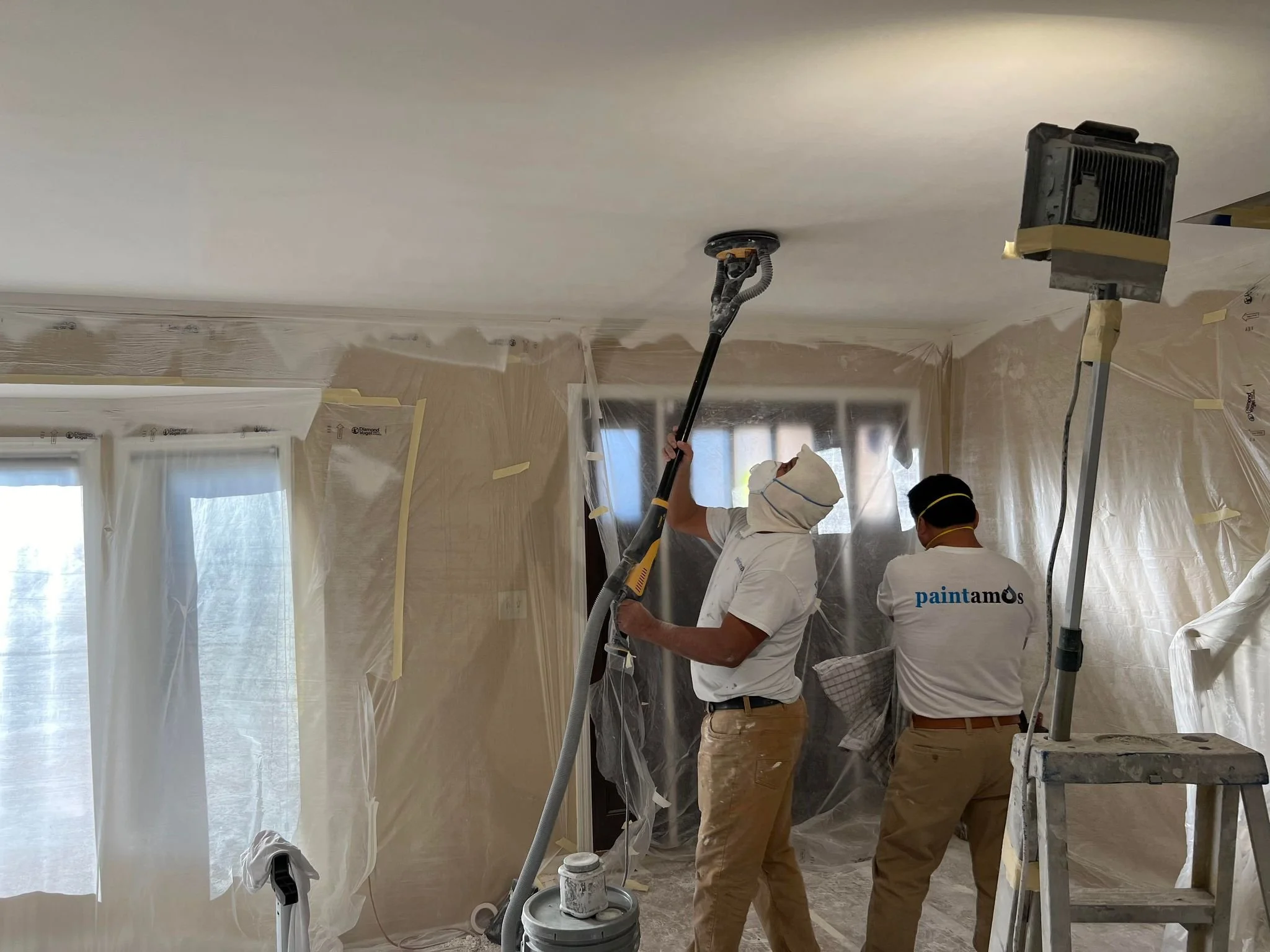 Two painters are painting a ceiling in a room covered with plastic sheeting. One is using a spray gun, and the other is holding a cloth, both wearing beige pants and white shirts, with one shirt displaying a logo.