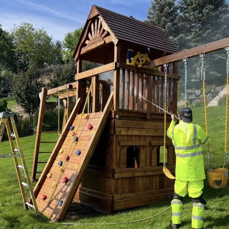 Worker cleaning a large wooden outdoor playhouse with swings, a climbing wall, and a slide in a grassy backyard.