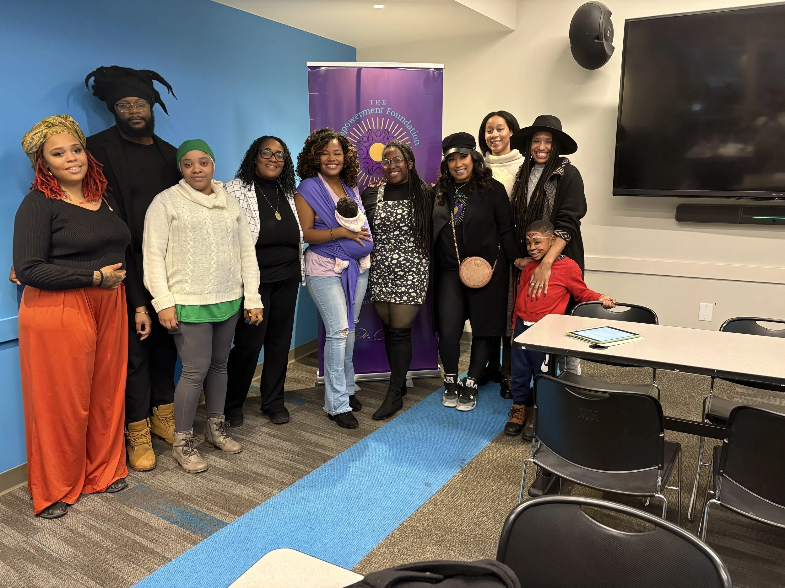 A diverse group of ten people, including men, women, and children, standing indoors in front of a purple banner that reads 'The Empowerment Foundation.' They are smiling and pose for a group photo in a room with a TV and tables.