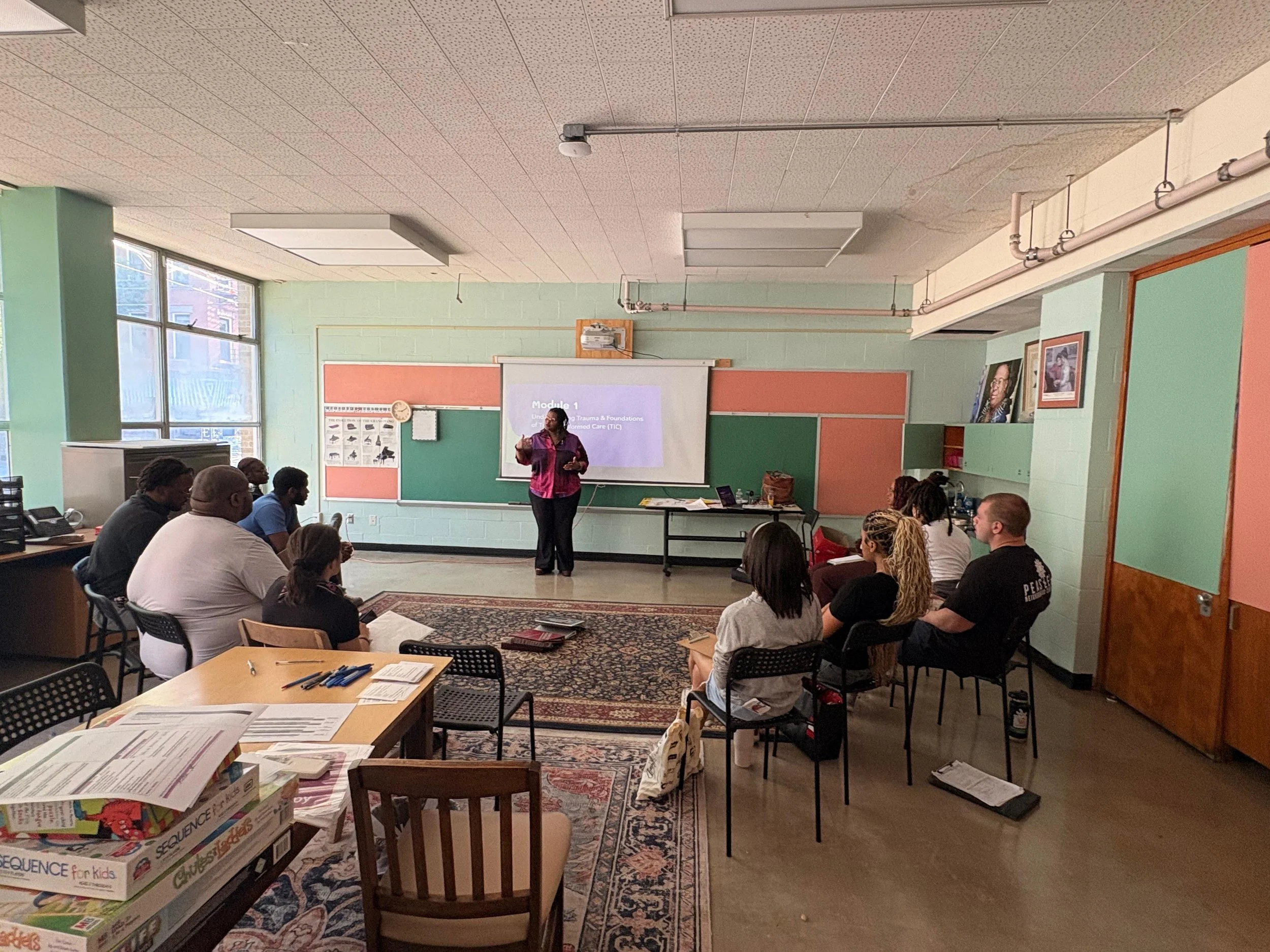 Classroom scene with a woman standing at the front giving a presentation, students seated and listening, colorful walls, large windows, and educational posters.