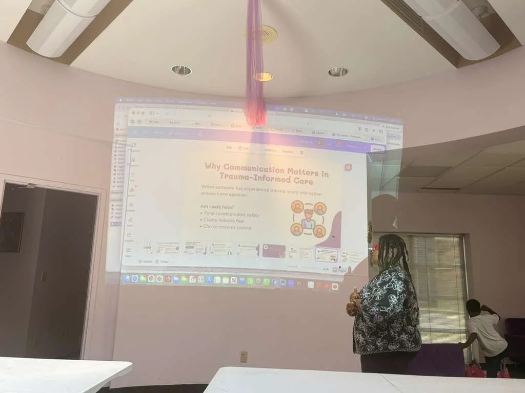 A woman stands in a room with pink walls, giving a presentation projected on the wall titled "Why Communication Matters in Trauma-Informed Care". Another person is seated at a table and appears to be listening lightly.