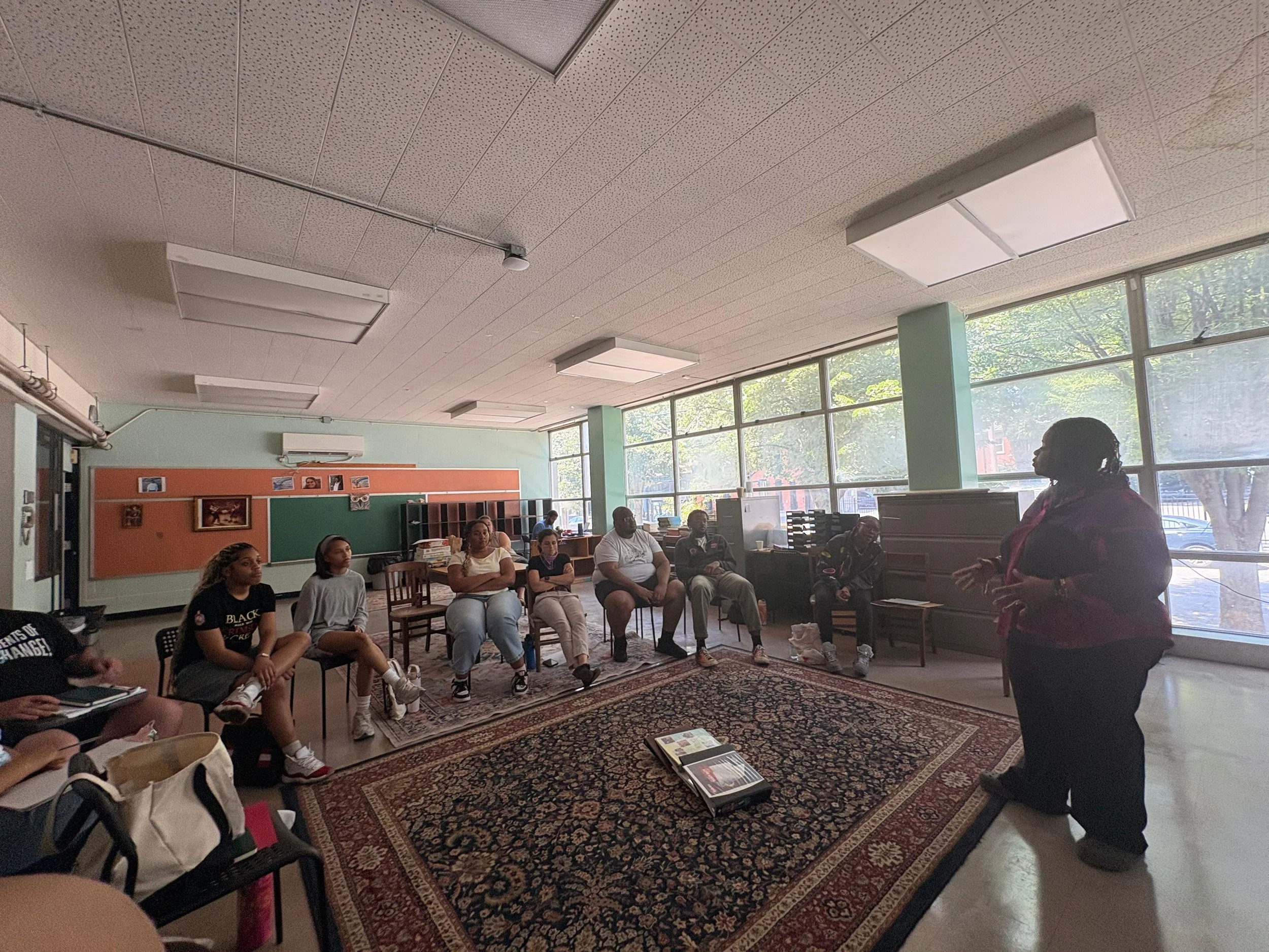 A group of diverse students sitting in a classroom, listening to a woman standing and speaking at the front of the room.