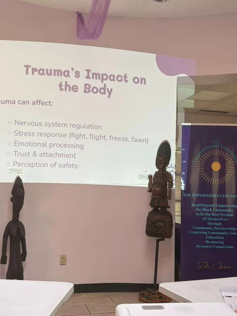 A presentation slide titled "Trauma's Impact on the Body" listing effects of trauma including nervous system regulation, stress response, emotional processing, trust and attachment, and perception of safety. The room has display statues and a banner from The Empowerment Foundation.
