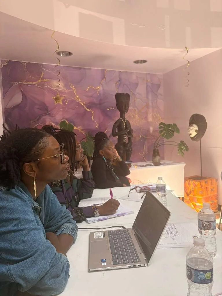 Group of women seated at a table in a room with purple and gold decor, listening attentively during a meeting or workshop, with notebooks, pens, and water bottles on the table.