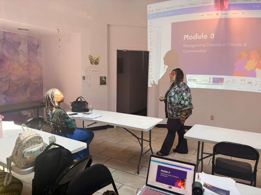 A woman giving a presentation in a classroom or training room, with a projected slide titled 'Module 3: Recognizing Trauma in Clients & Communities,' while a woman with gray dreadlocks and glasses listens. There are tables, chairs, and backpacks in the room.