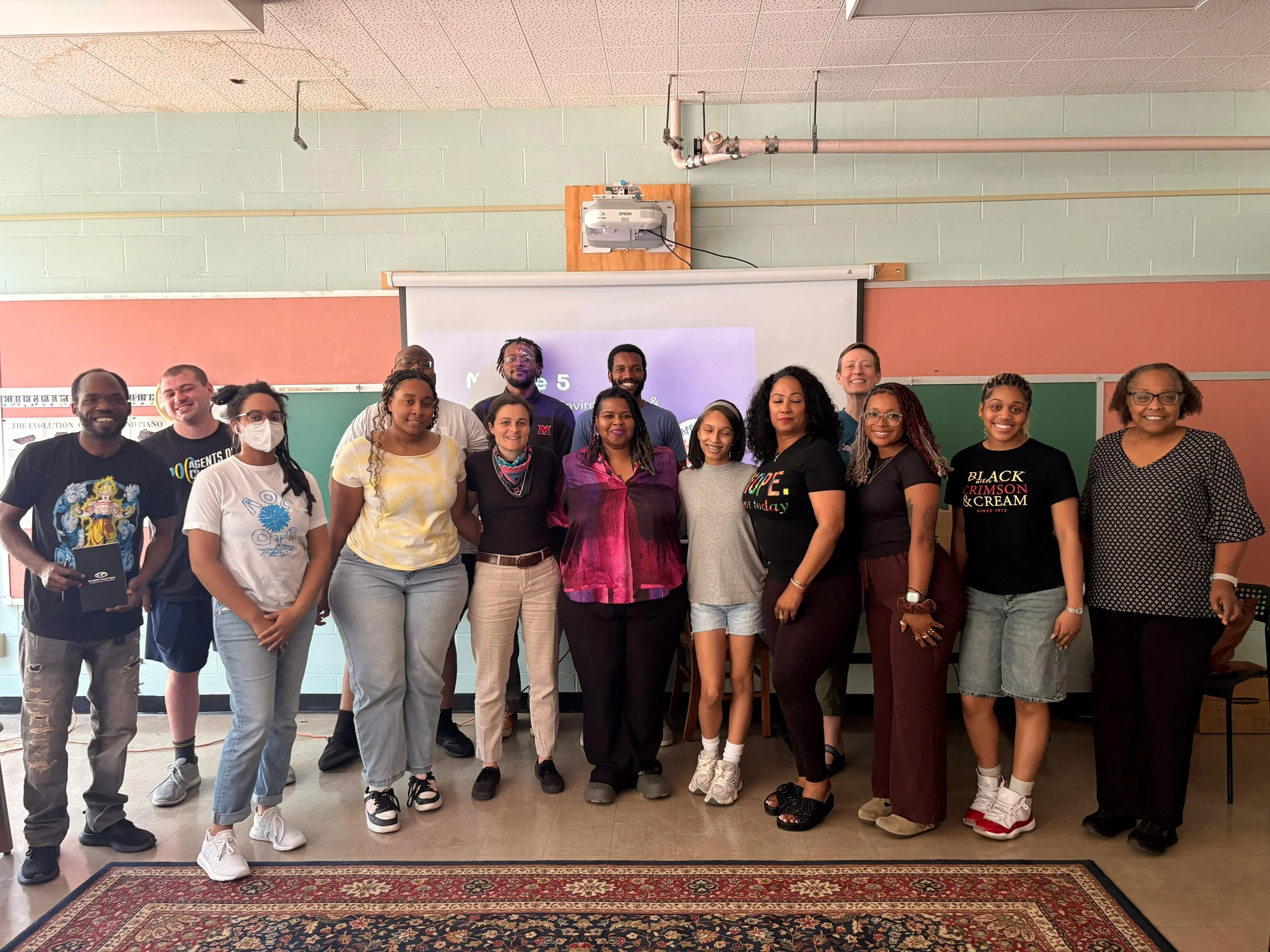 Group of diverse people standing in a classroom or auditorium, posing for a photo, with a projected presentation in the background.