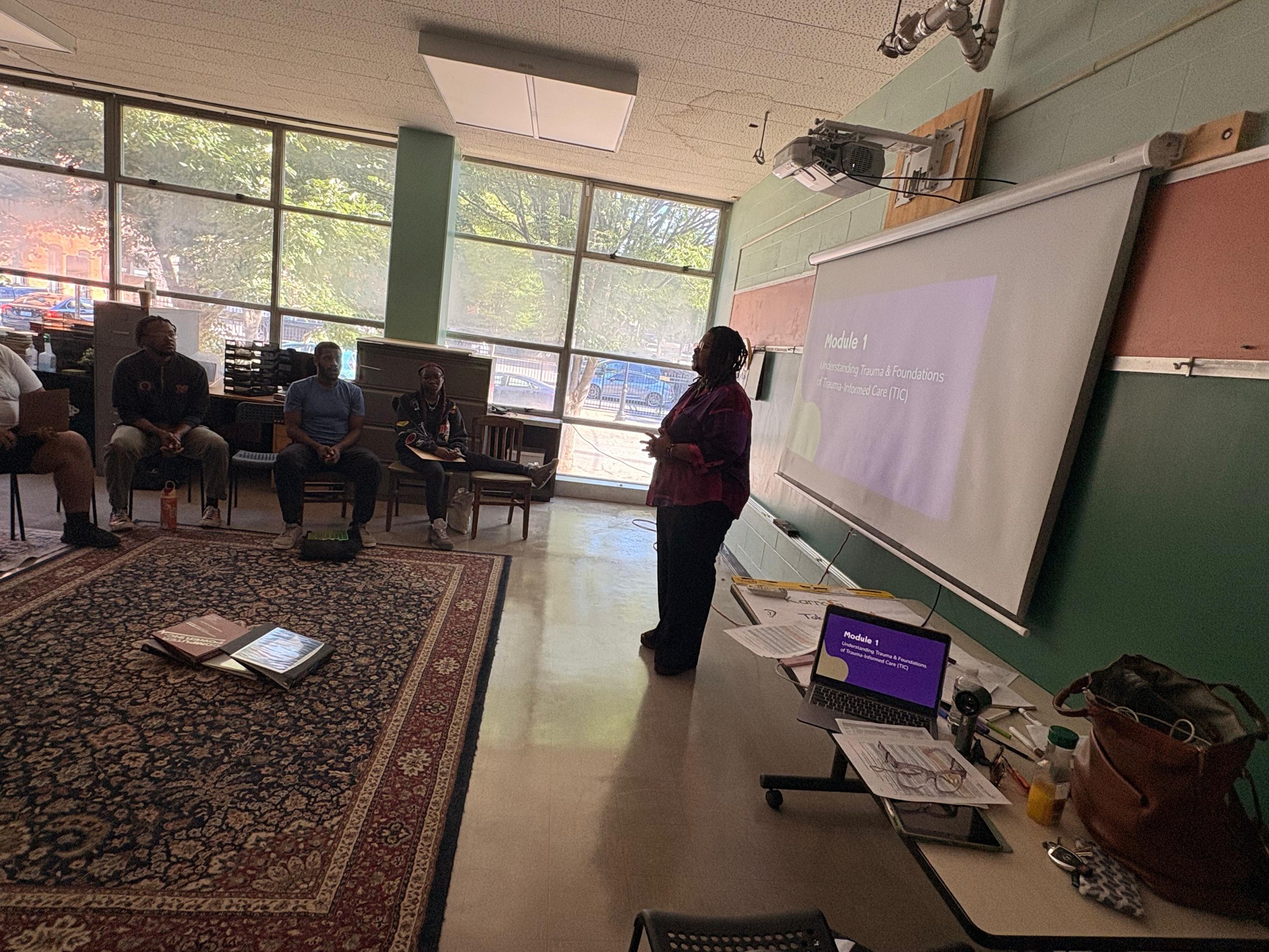 A classroom or training room with people seated in a semi-circle facing a woman speaker. There is a large screen displaying a presentation titled 'Module 1: Understanding Trauma & Foundations of Trauma-Informed Care (TIC).' Large windows let in natural light, and various tables and equipment are visible around the room.