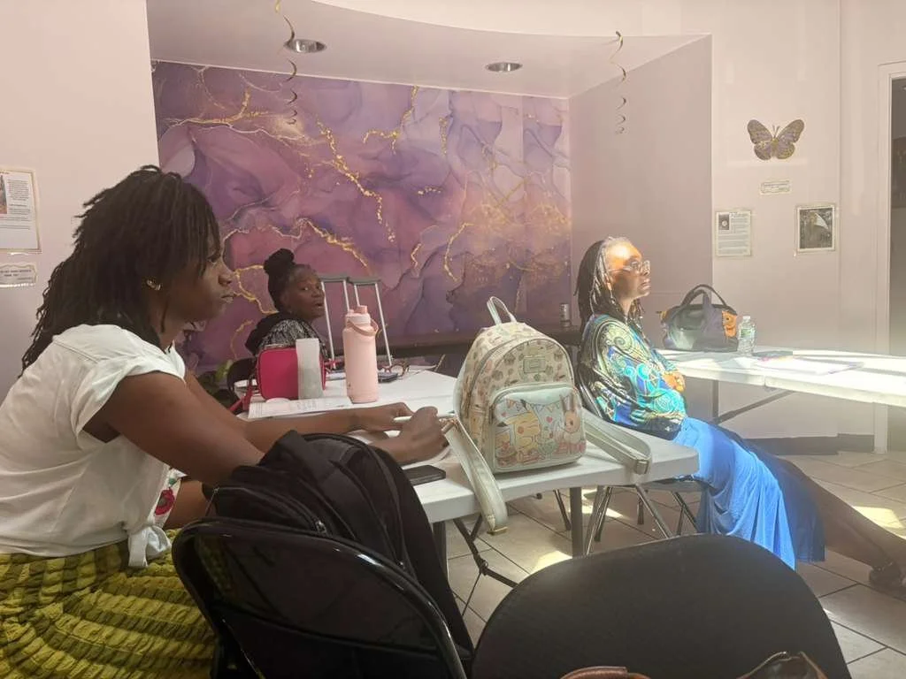 Three women sitting at tables in a room with purple marble wall, butterfly decoration, and framed pictures, engaged in a meeting or class.