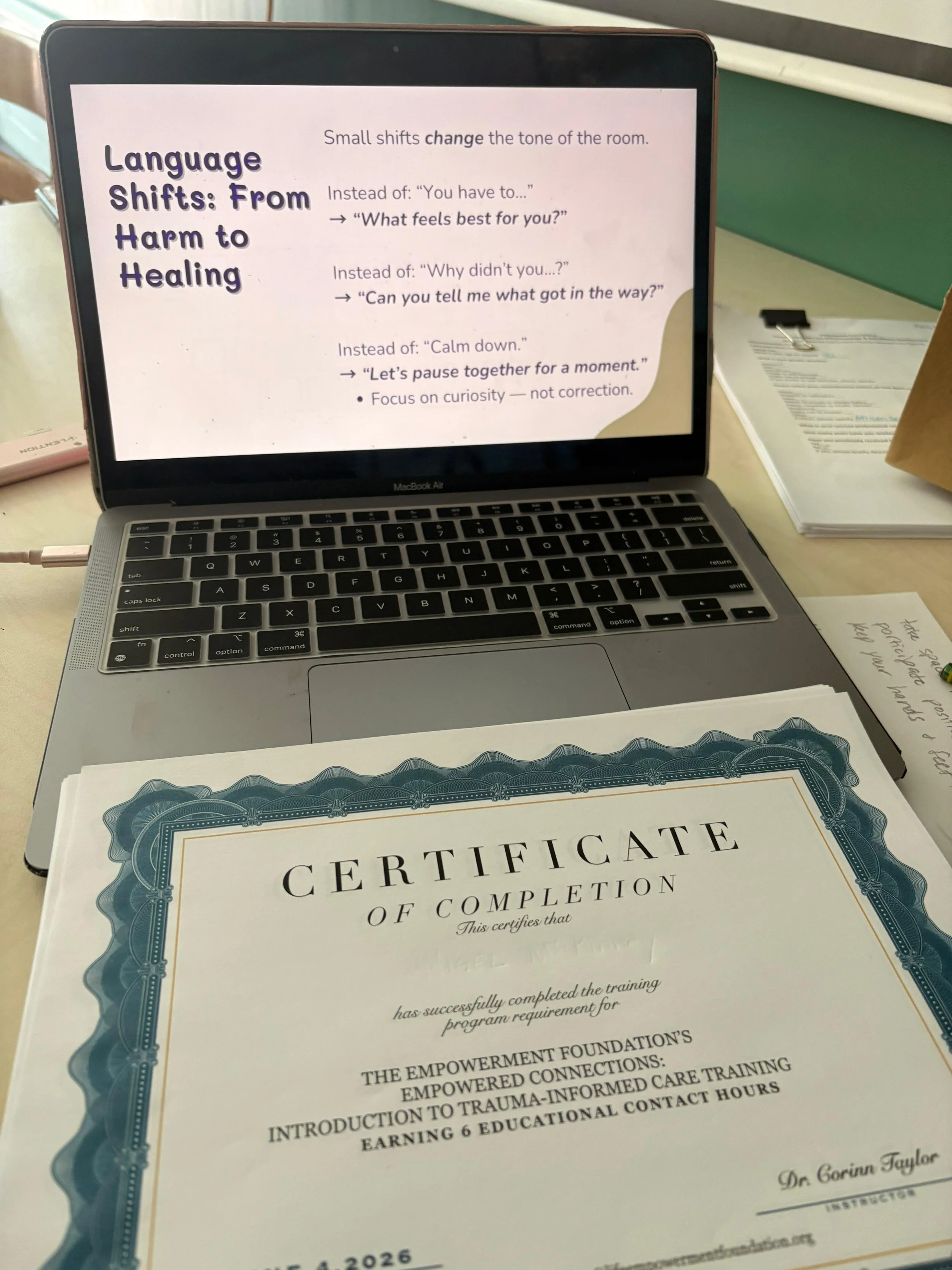 A laptop showing a presentation slide titled "Language Shifts: From Harm to Healing" with tips on changing phrases to promote healing. In front of the laptop, a certificate of completion is visible, partially covering the laptop. To the right, there are papers and a clipboard on the table.