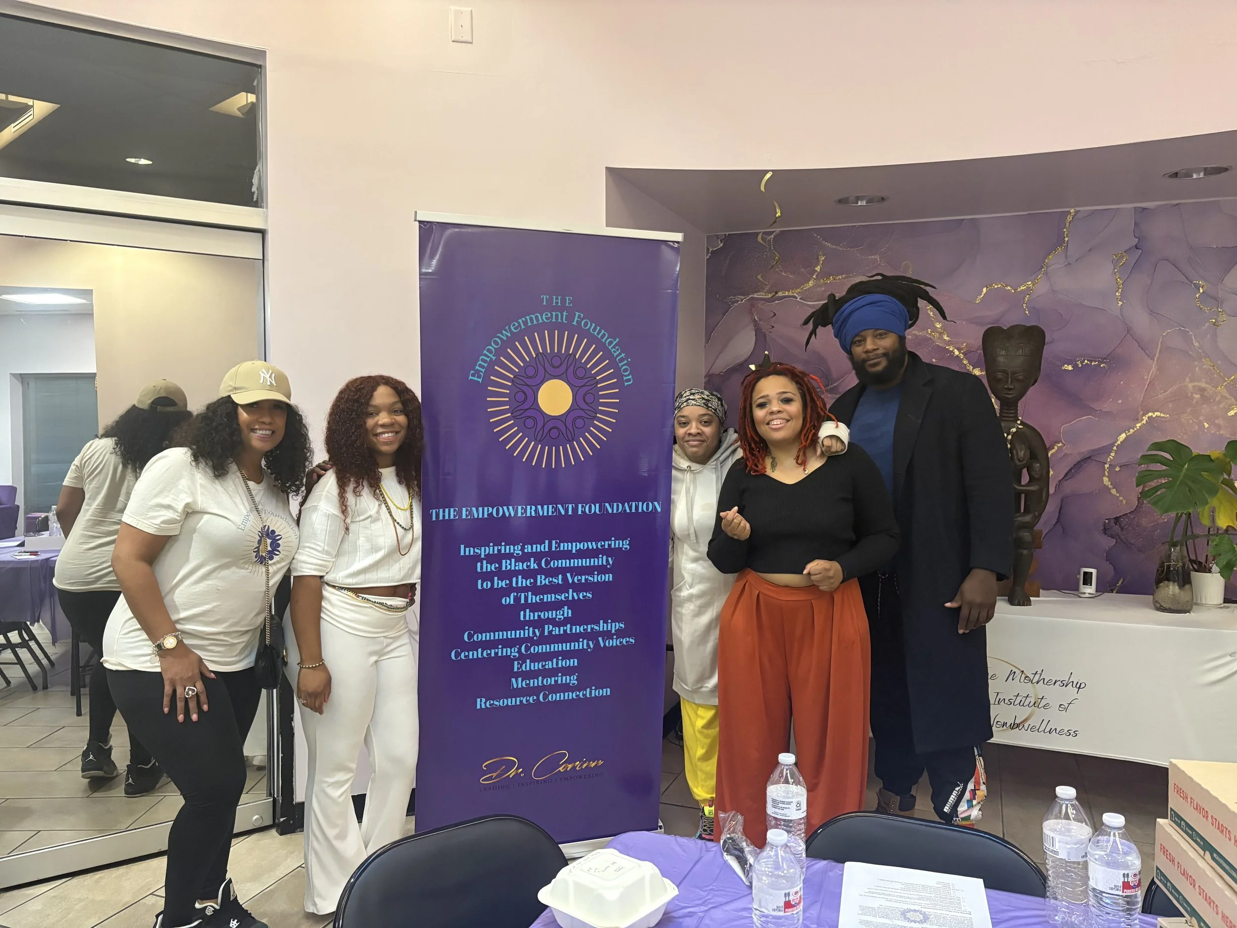 Group of six people standing indoors next to a large purple banner for The Empowerment Foundation, with a purple and gold painted wall and African sculpture in the background.