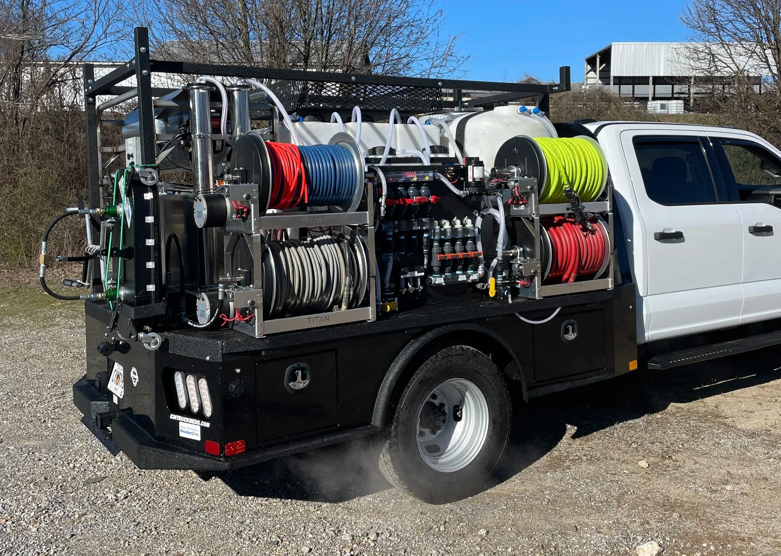 A fire truck equipped with various hoses and firefighting equipment, parked on a gravel surface with a clear blue sky and trees in the background.