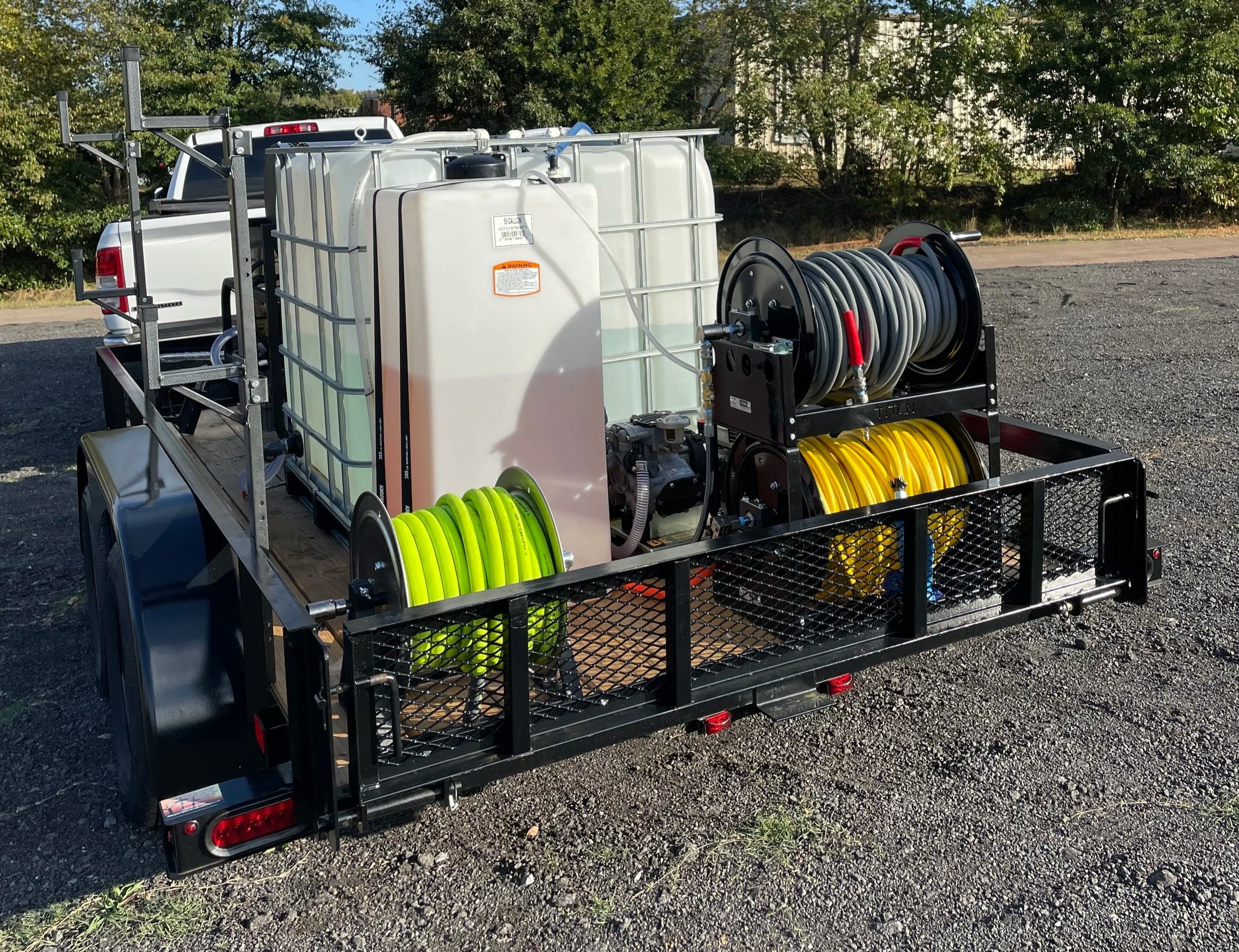 A mobile water hose and equipment trailer with large water tanks, a hose reel, and various hoses, parked on a gravel lot with trees in the background.